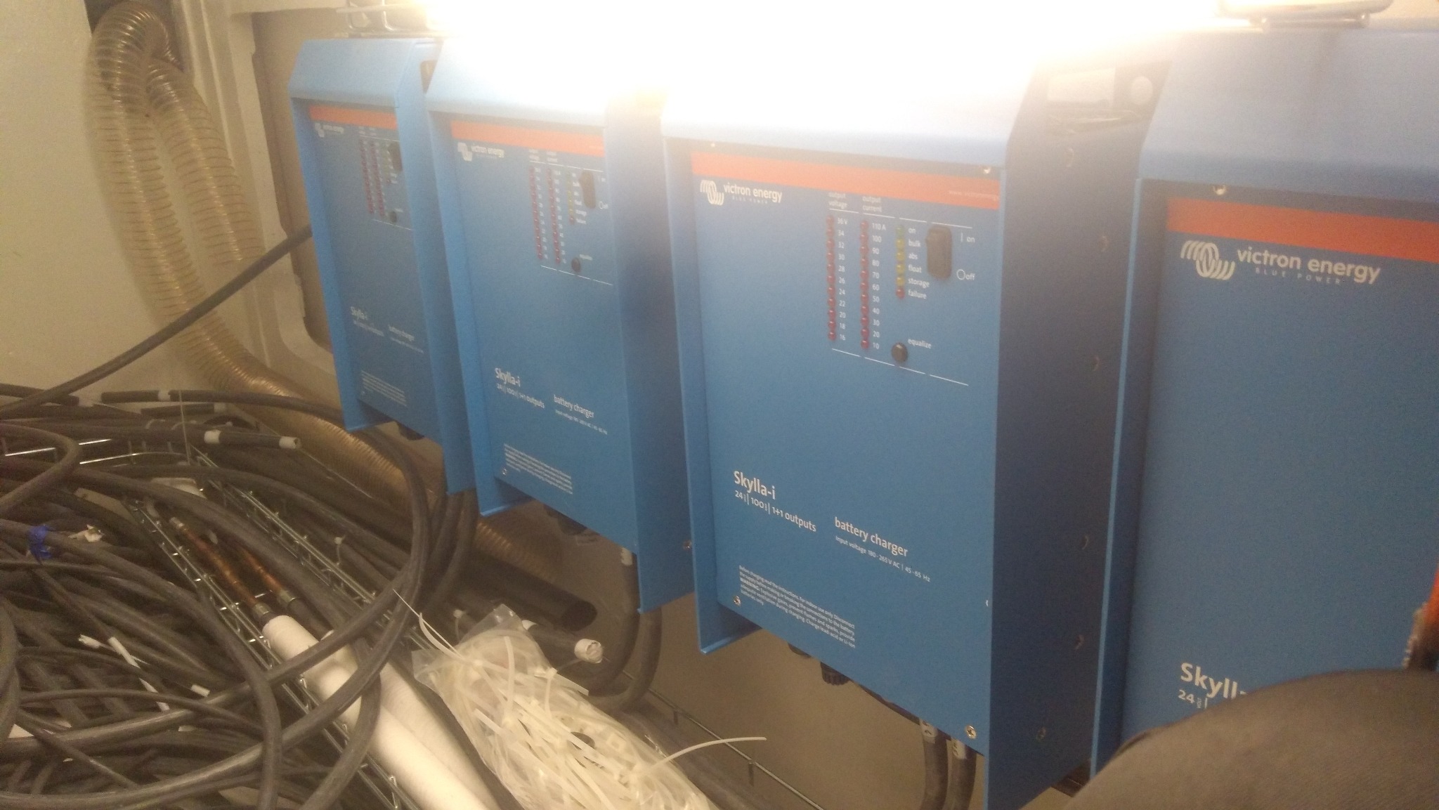 Cztery niebieskie ładowarki akumulatorów Victron Energy Skylla-i, z widocznymi kablami i przewodami w tle, zainstalowane w pomieszczeniu technicznym.