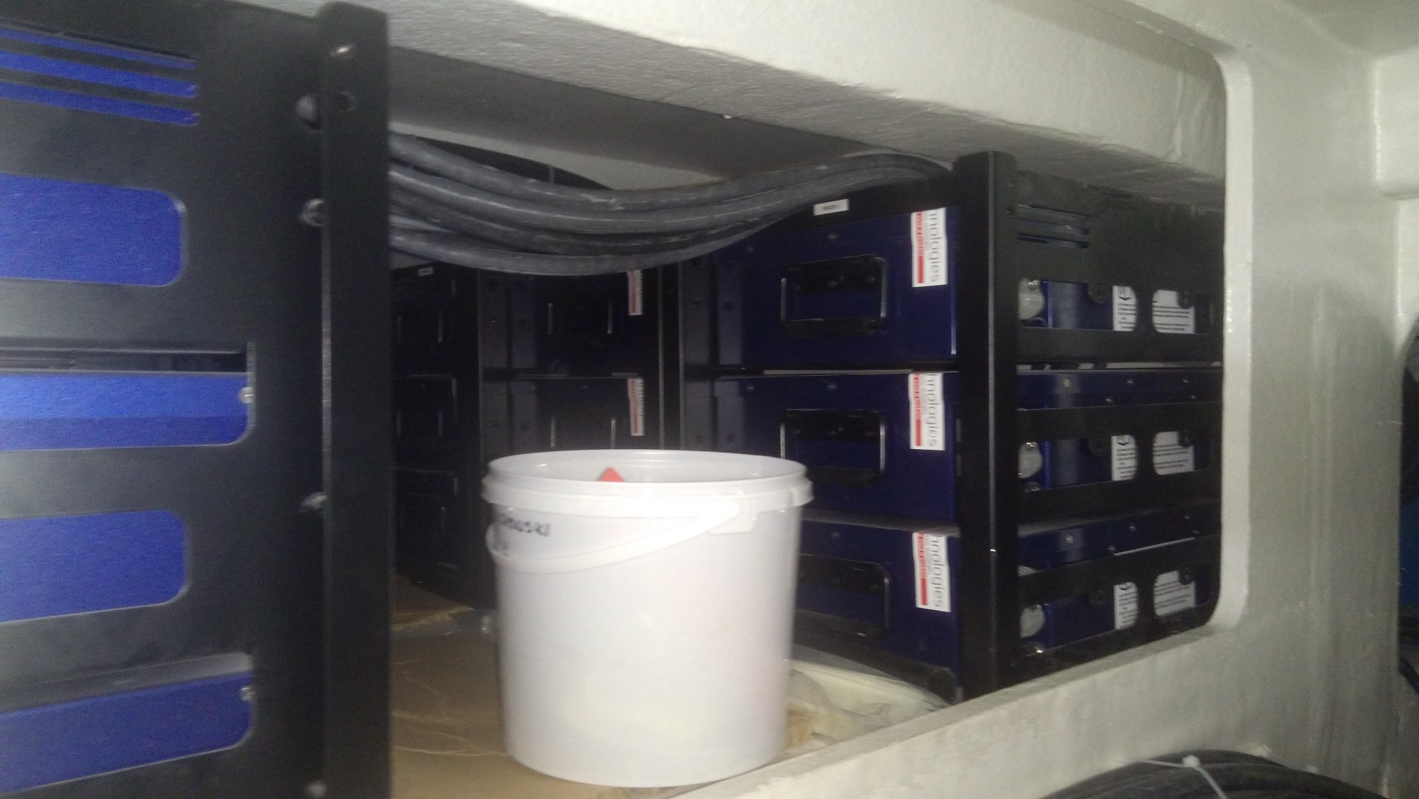 Rząd akumulatorów w szafie, widoczne kable i wiadro z farbą w pomieszczeniu.