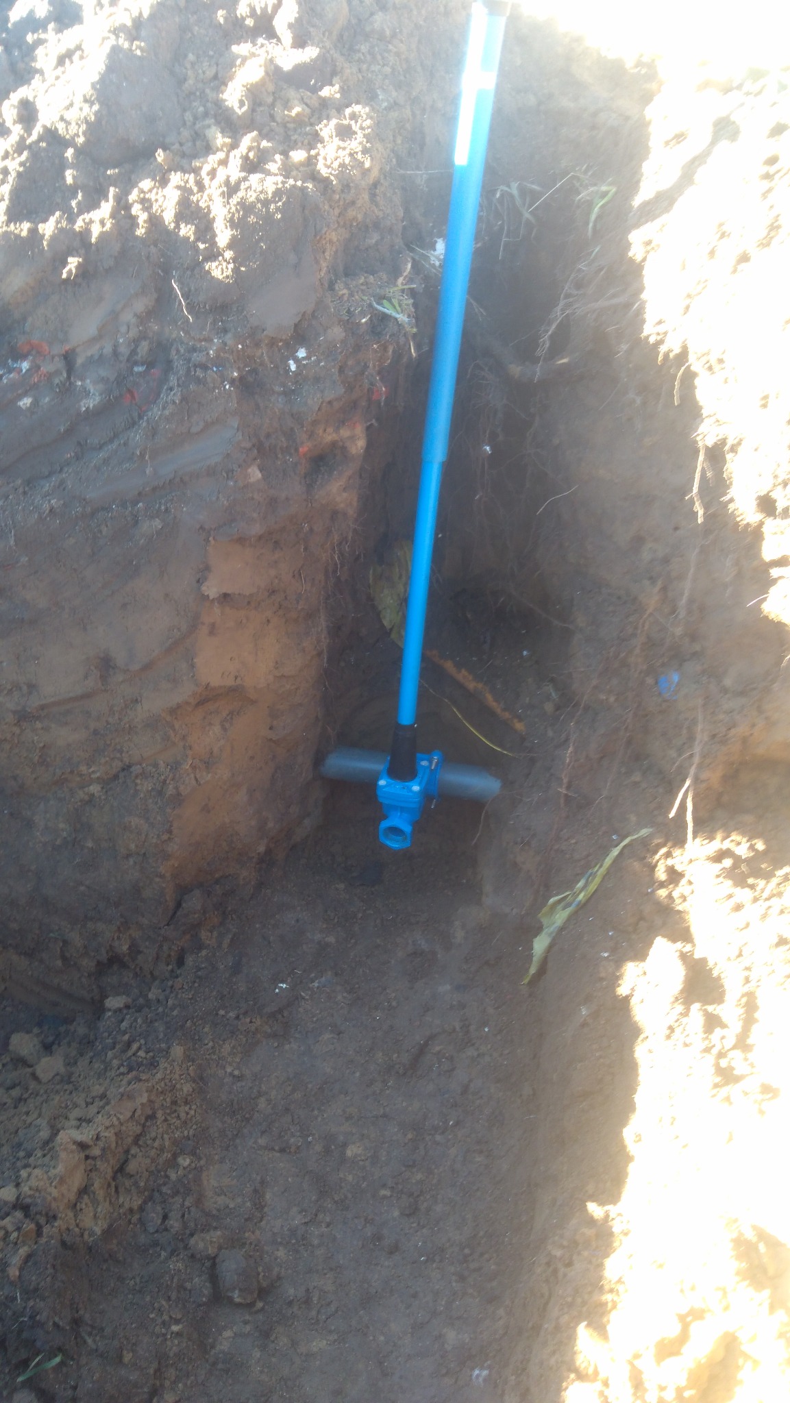 Niebieski zawór trójdrożny z rurą w wykopie ziemnym, widoczne warstwy gleby i korzenie.