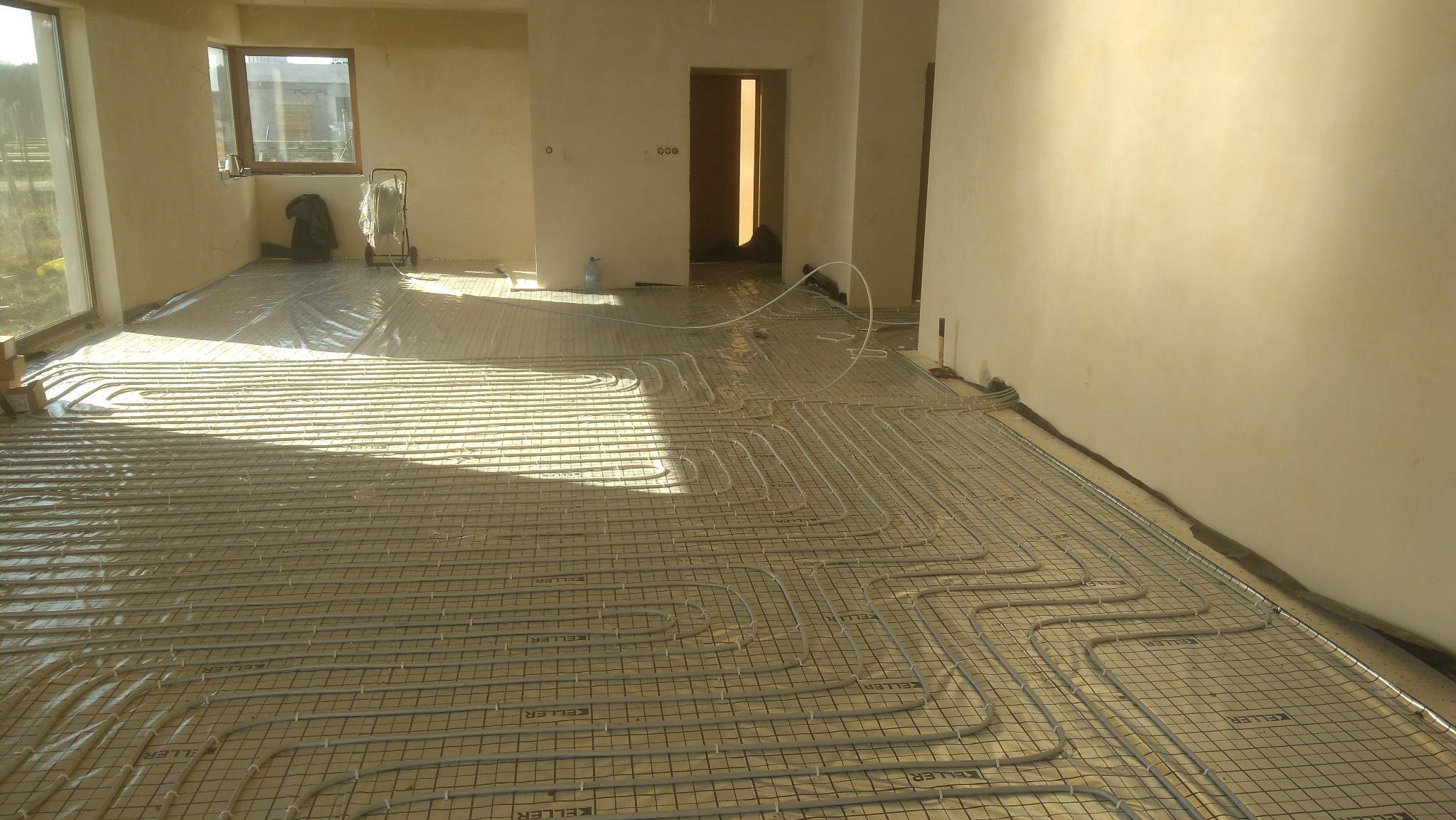 Rozłożona instalacja ogrzewania podłogowego na siatce w surowym wnętrzu, widoczne rury i folia ochronna.