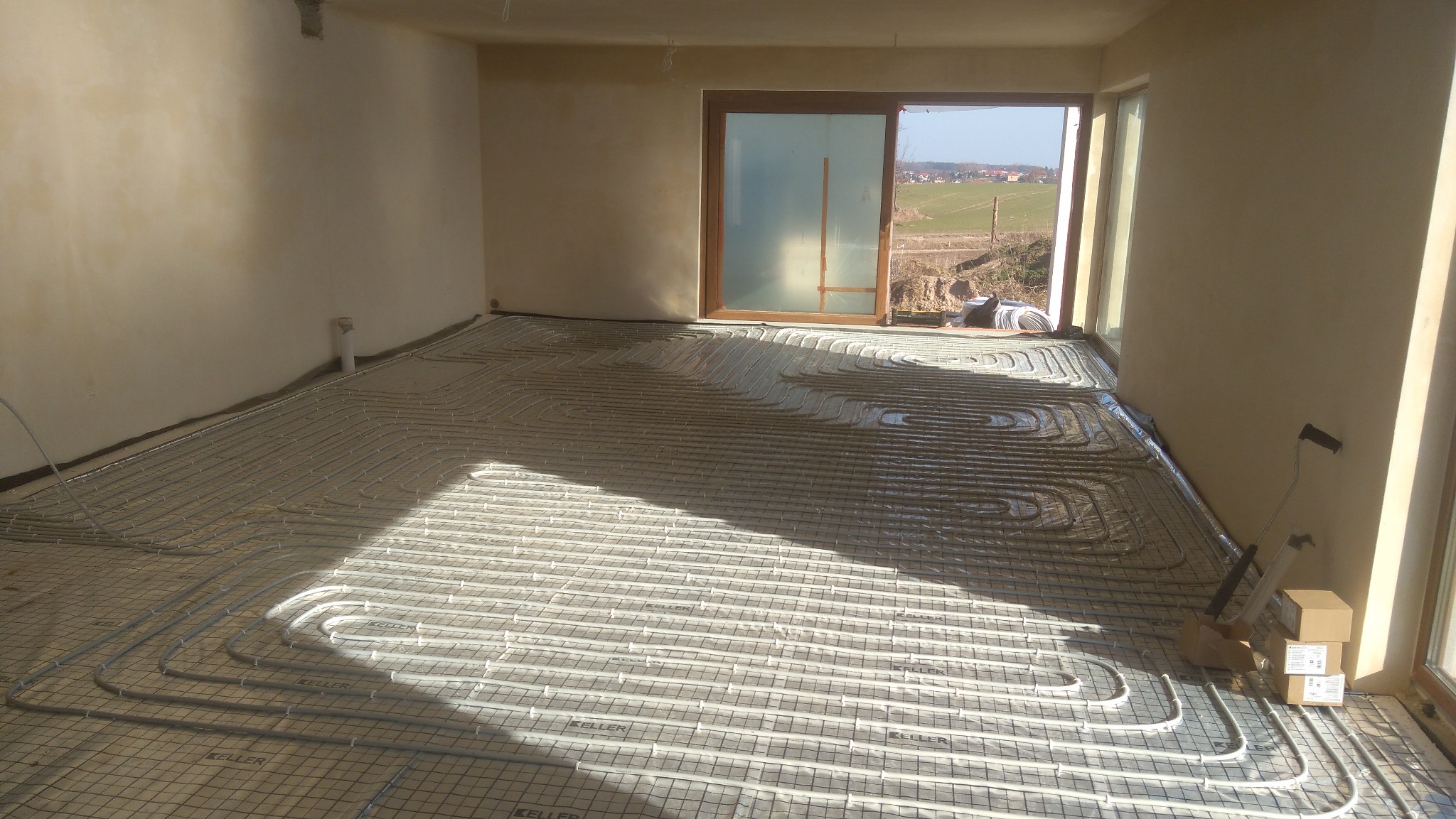 Rozłożone rury ogrzewania podłogowego na siatce montażowej w surowym wnętrzu z widokiem na krajobraz za oknem.