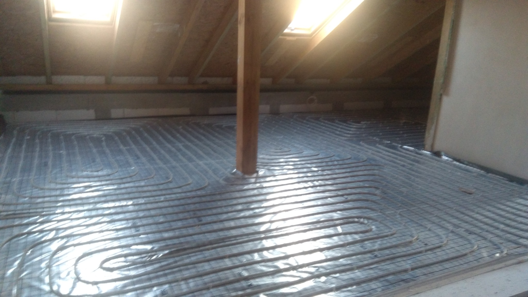 Układanie rur ogrzewania podłogowego na poddaszu, widoczne okno dachowe i drewniany słup podtrzymujący konstrukcję dachu.