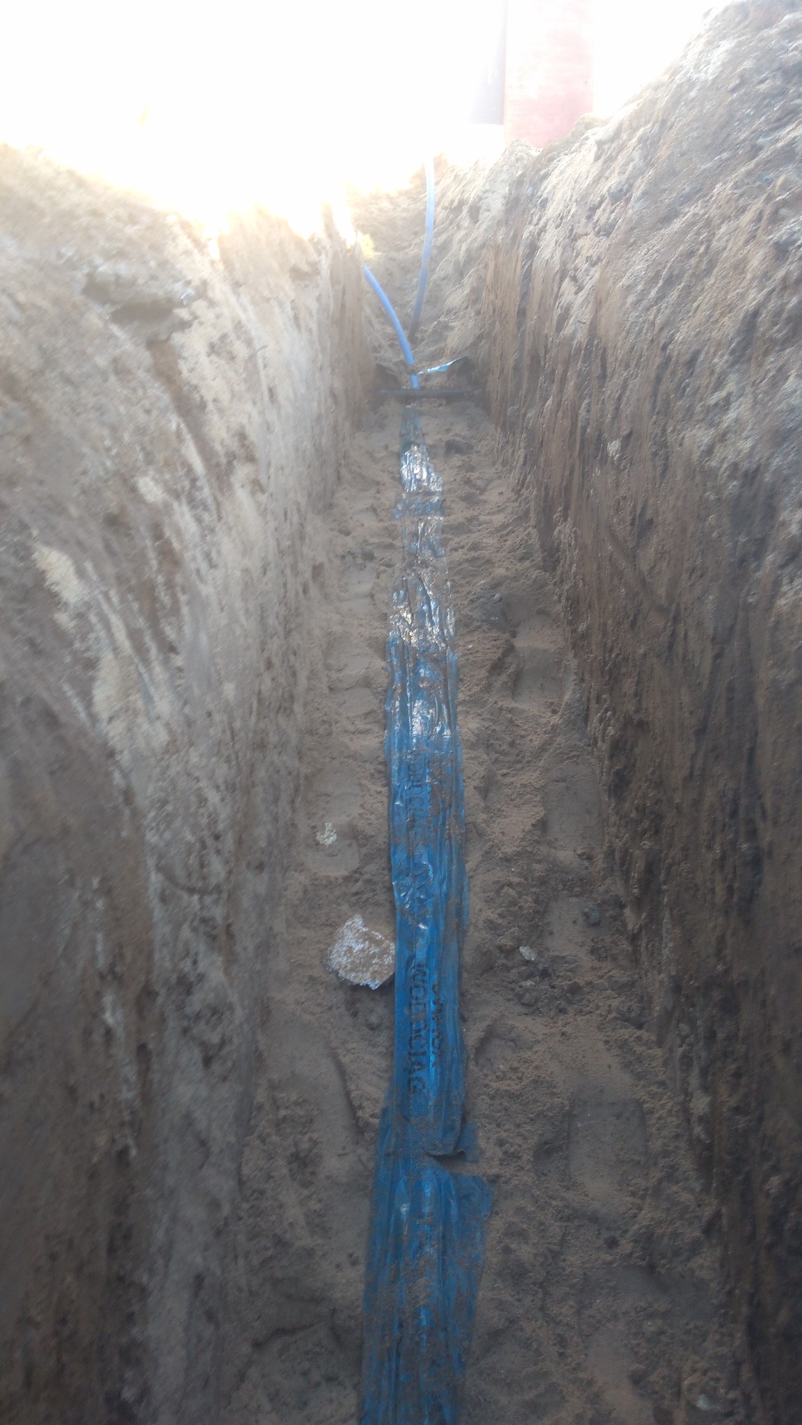 Wykop w ziemi z niebieską rurą osłonową i fragmentami niebieskich rurek. Widoczny piasek i ziemia.