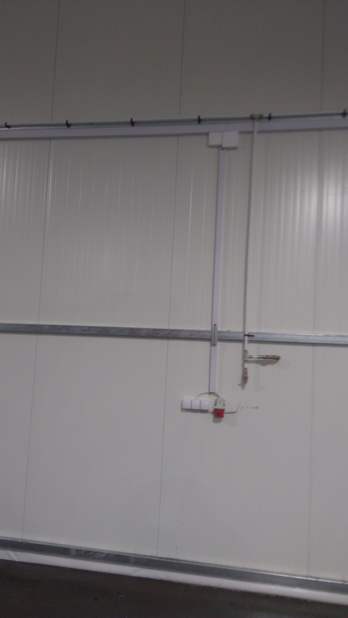 Biała ściana z panelami, widoczne metalowe szyny montażowe, biała rurka z kablami i gniazdami elektrycznymi, oraz rurka z zaworem.