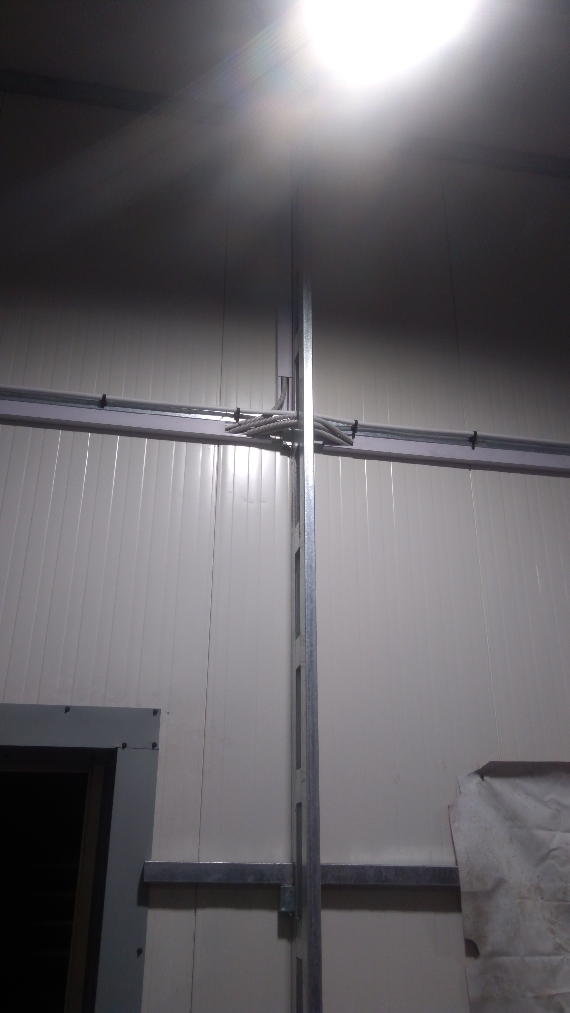 Wnętrze hali produkcyjnej z widocznymi kablami w peszlach prowadzonymi po ścianie i metalową konstrukcją wsporczą.