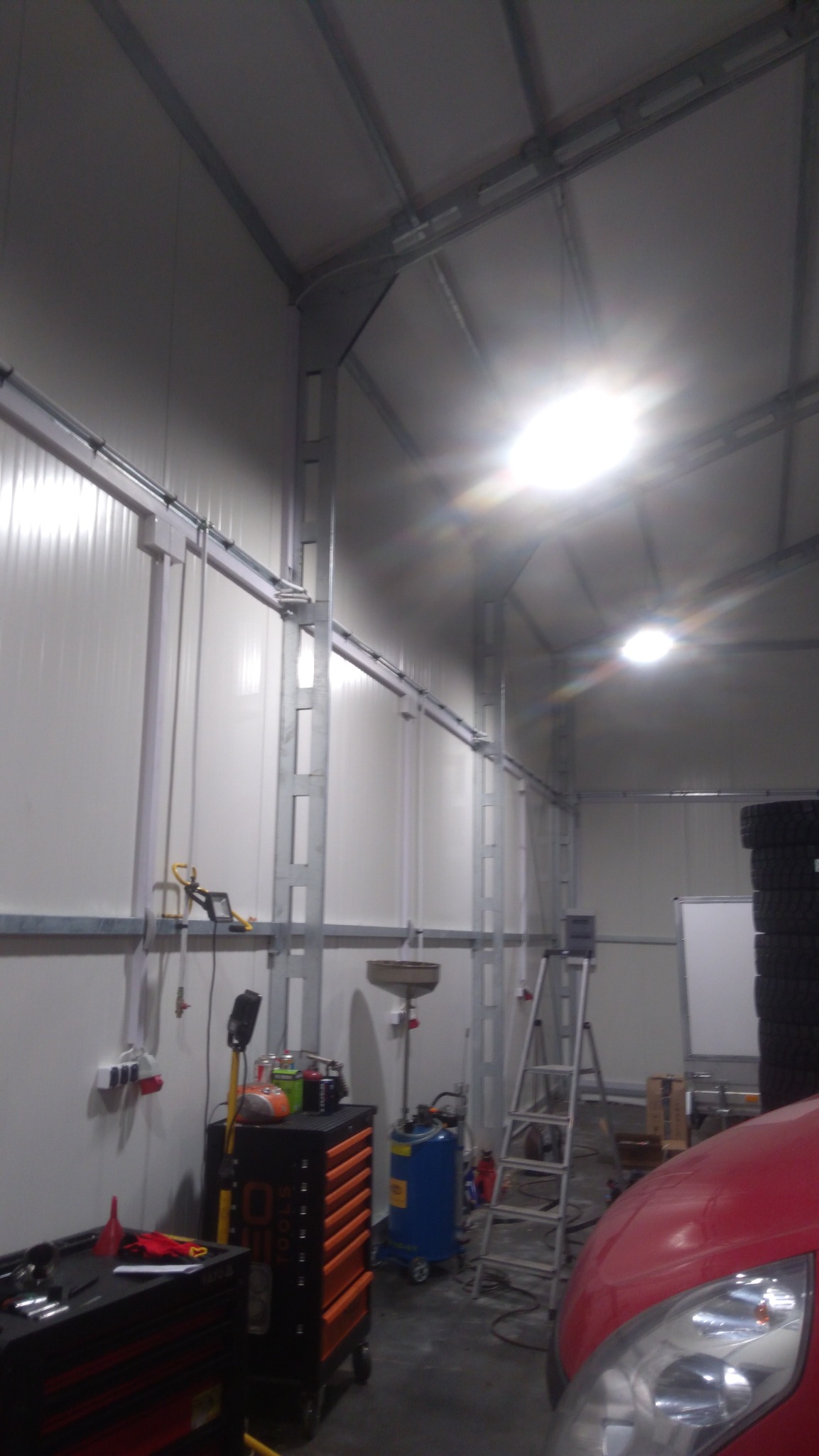 Wnętrze warsztatu z metalową konstrukcją stropu, narzędziami na wózku i oponami w tle, oświetlone lampą halogenową.