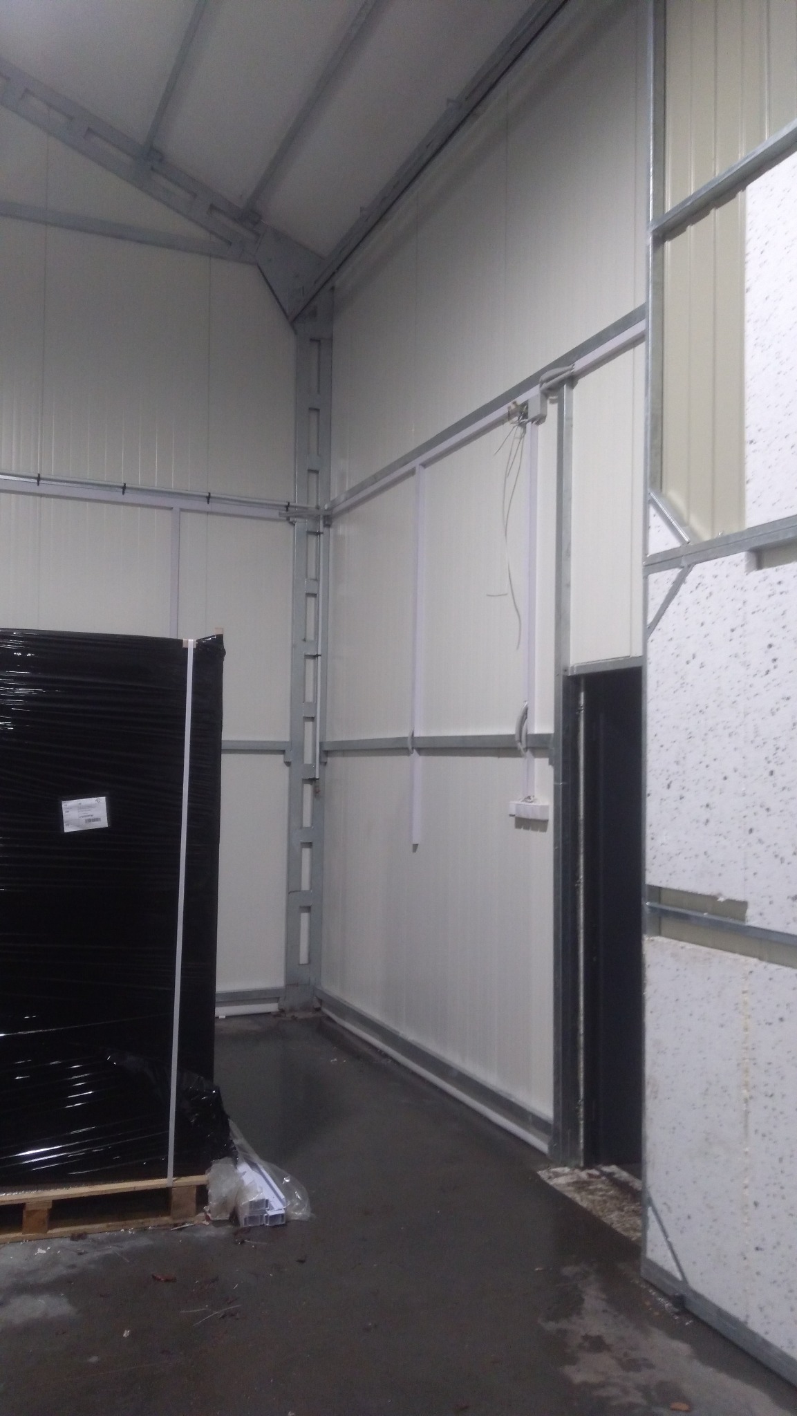 Wnętrze hali z metalową konstrukcją i panelowymi ścianami, widoczne przewody elektryczne wystające ze ściany oraz paleta z zapakowanym towarem w czarnej folii.