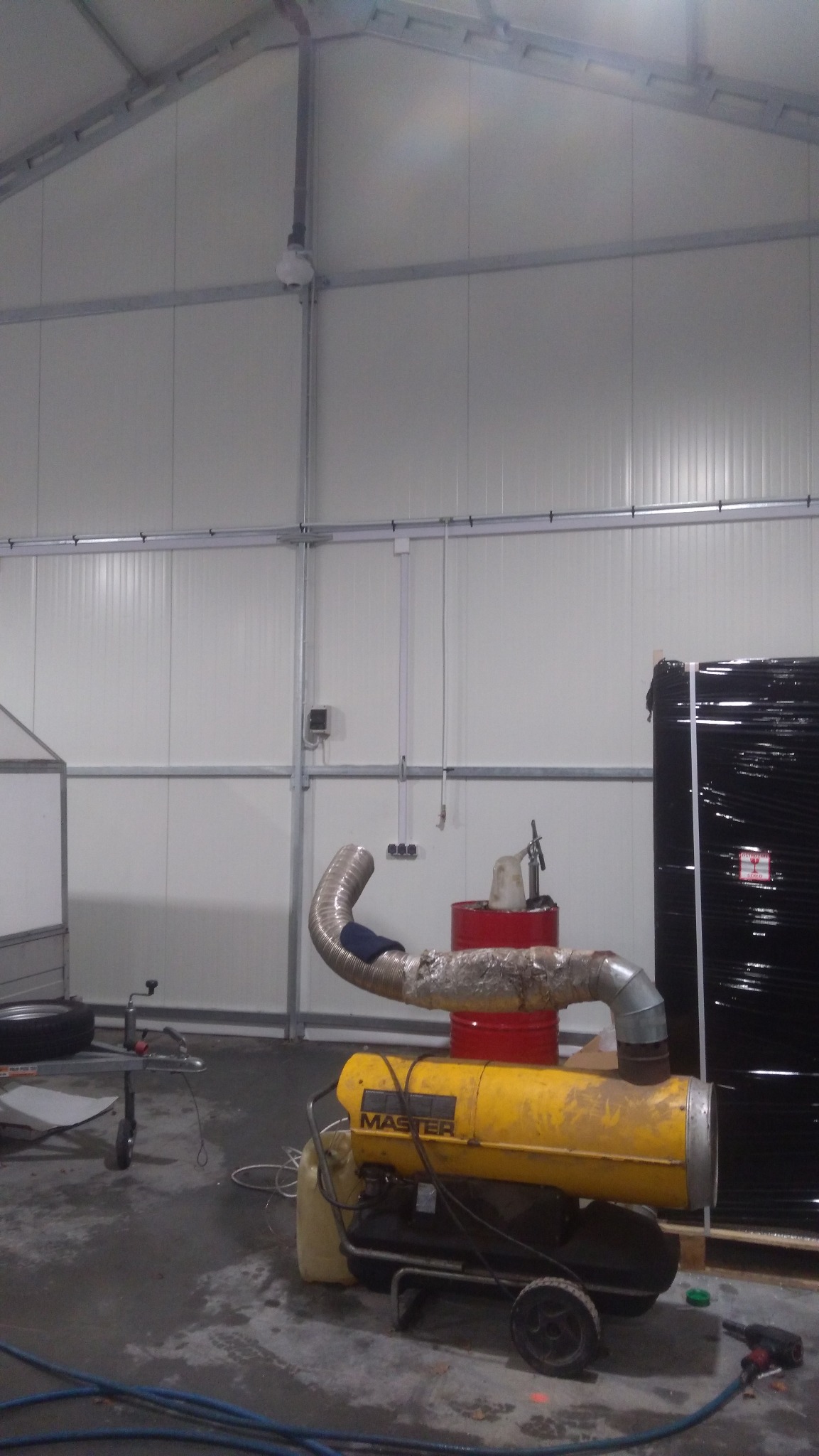 Przenośny nagrzewnica olejowa MASTER ustawiona wewnątrz hali z widoczną instalacją elektryczną pod sufitem i przewodami na ścianie.