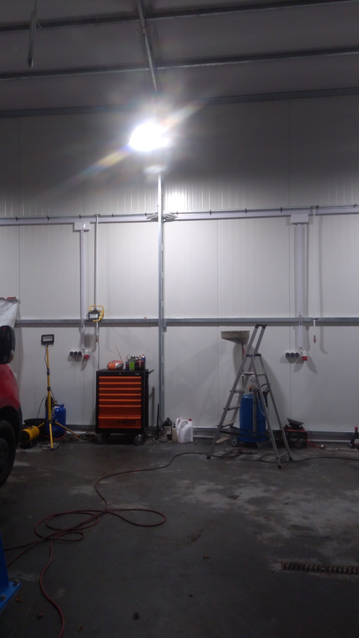 Wnętrze warsztatu z narzędziami i sprzętem: szafka narzędziowa NEO, drabina, lampa robocza i rozłożone kable na betonowej posadzce, oświetlone jasnym światłem górnym.