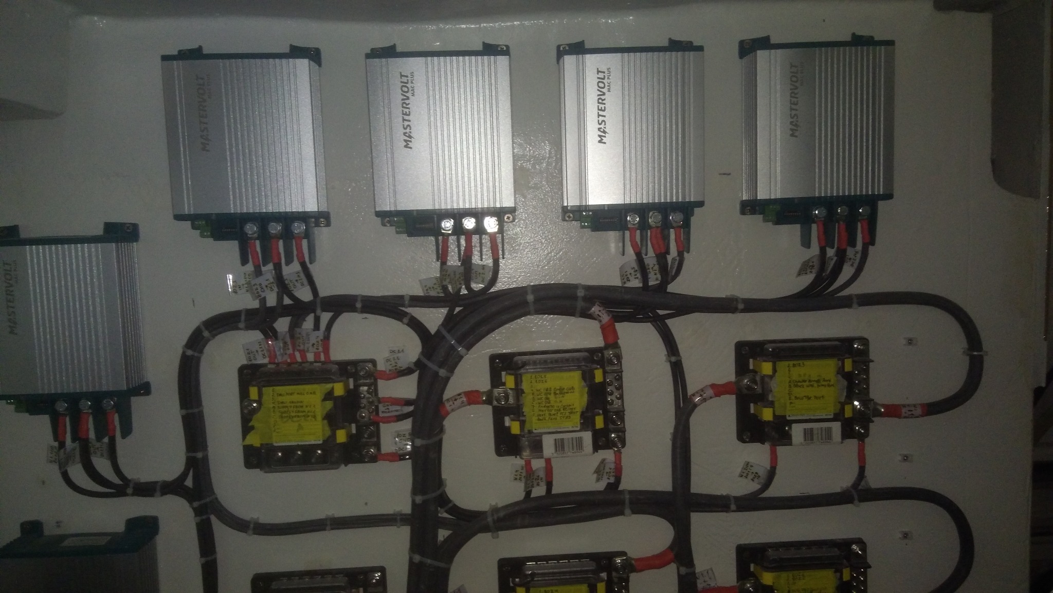 Panel z zamontowanymi srebrnymi urządzeniami Mastervolt z podłączonymi kablami, widoczne również transformatory z żółtymi etykietami, całość zamontowana na białej ścianie.