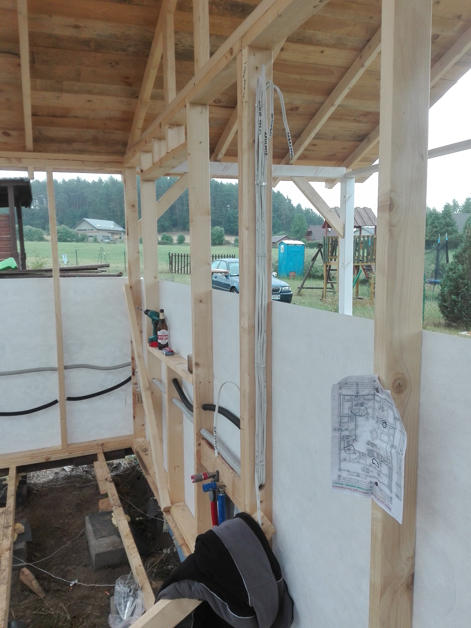 Wnętrze szkieletowej konstrukcji budynku w trakcie budowy, widoczne instalacje: elektryczne w peszlach i wodno-kanalizacyjne, narzędzia, plan instalacji przytwierdzony do ściany, butelka piwa...