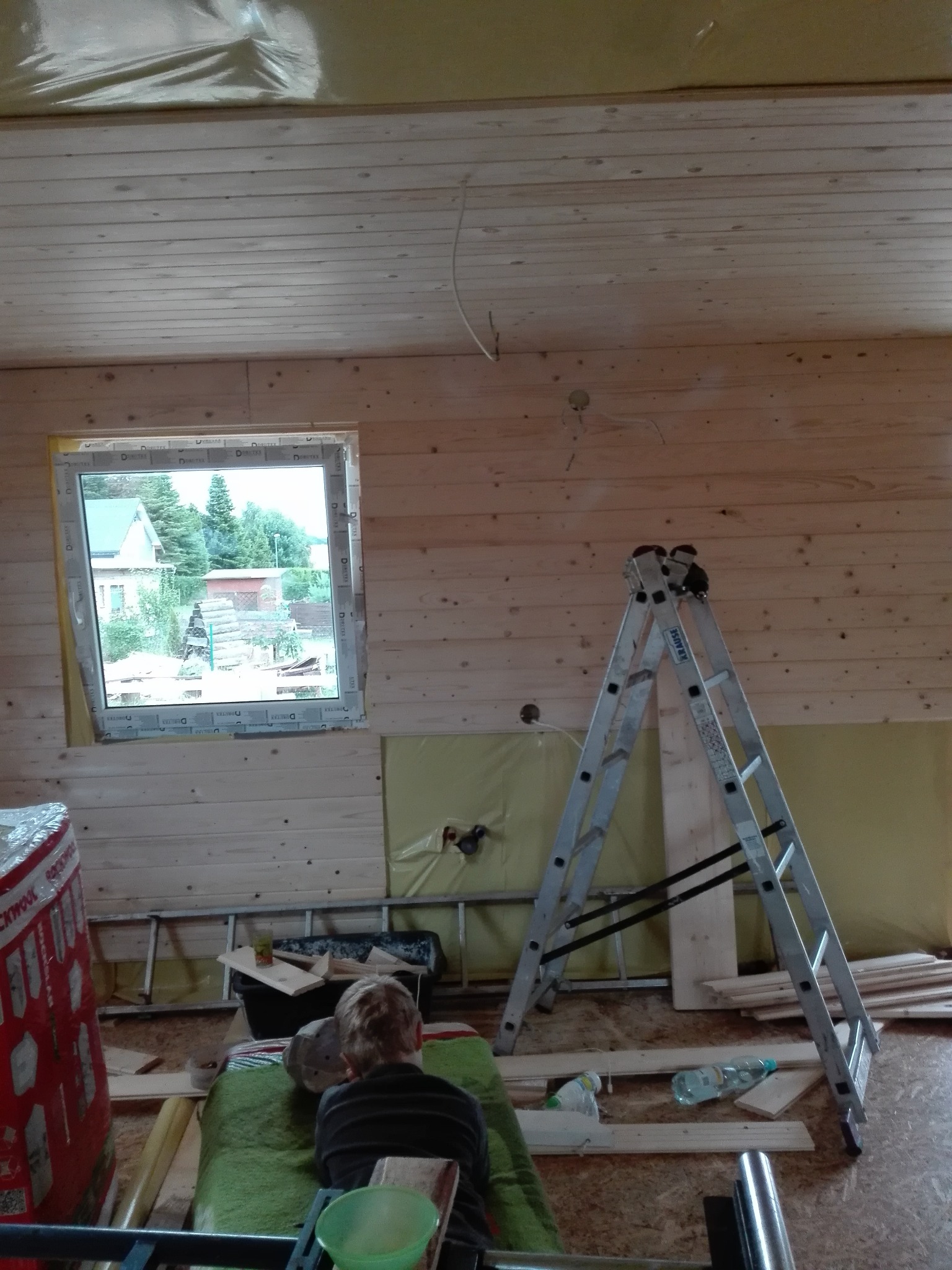 Wnętrze drewnianego domu w trakcie prac wykończeniowych: widoczne drewniane ściany i sufit, okno z widokiem na zewnątrz, drabina aluminiowa oparta o ścianę, materiały budowlane i narzędzia...