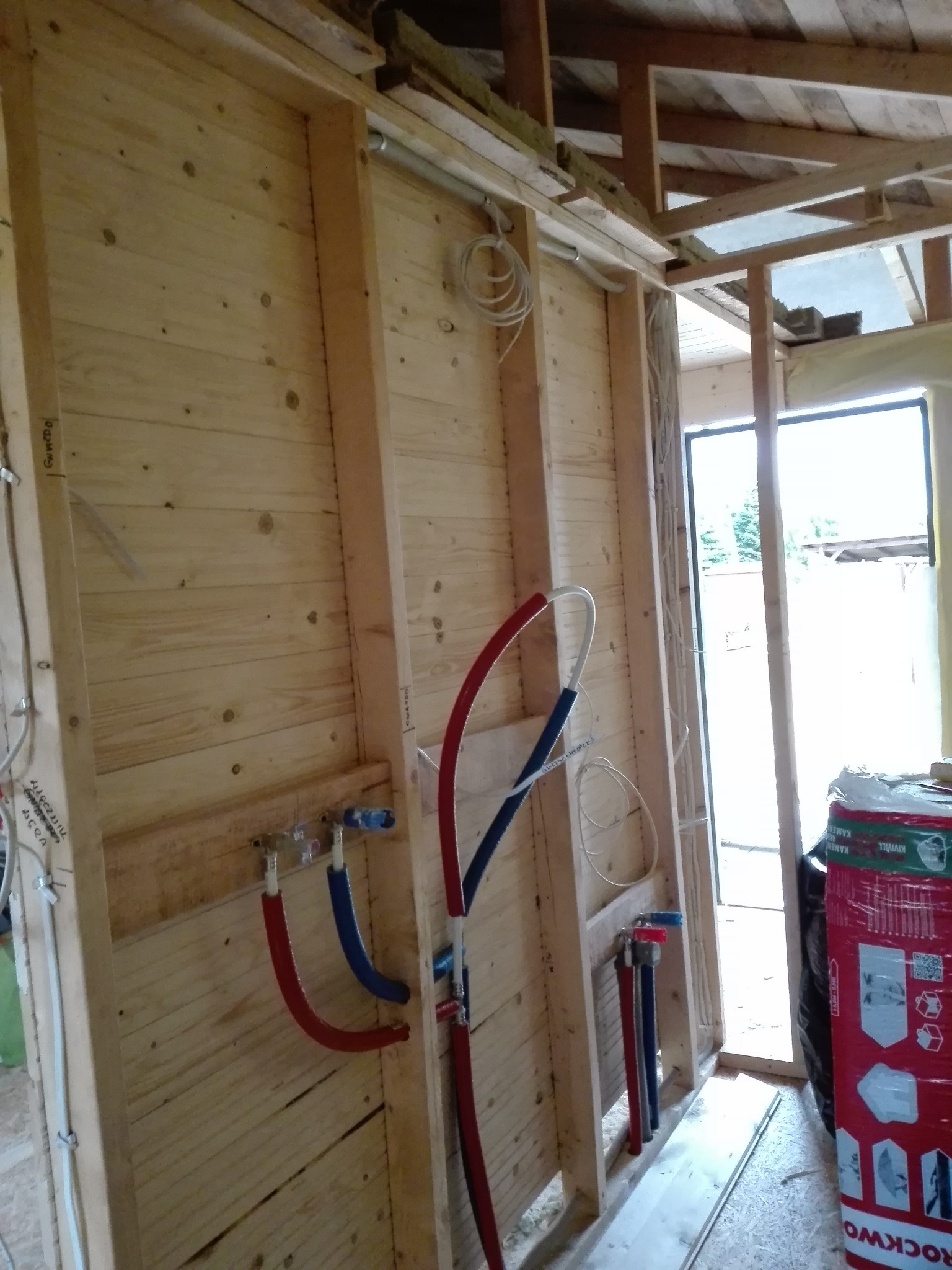 Surowa drewniana konstrukcja ściany z widocznymi rurami instalacji wodnej w kolorze czerwonym i niebieskim, przewody elektryczne w peszlach i częściowo ocieplony strop.