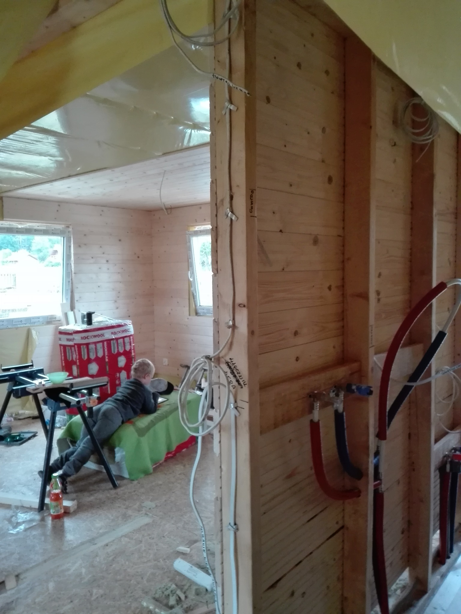 Wnętrze domu w trakcie remontu: widoczne drewniane ściany w trakcie budowy, instalacje elektryczne prowadzone po ścianach, rury instalacji wodnej oraz dziecko leżące na prowizorycznym łóżku...