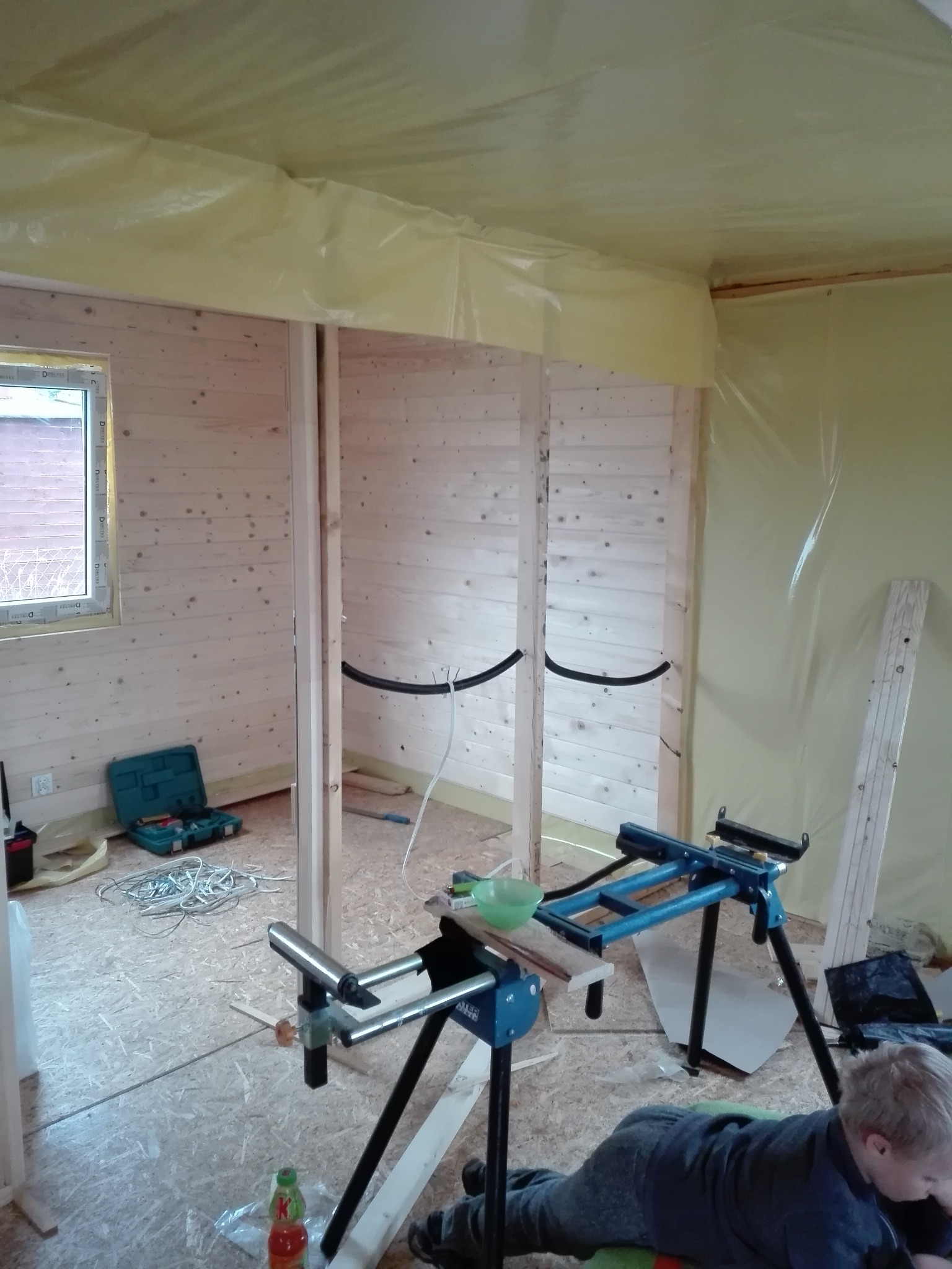 Wnętrze drewnianego domu w trakcie budowy w Rumi. Widoczne ściany z boazerii, instalacja elektryczna w peszlu, narzędzia i osoba pracująca na podłodze.