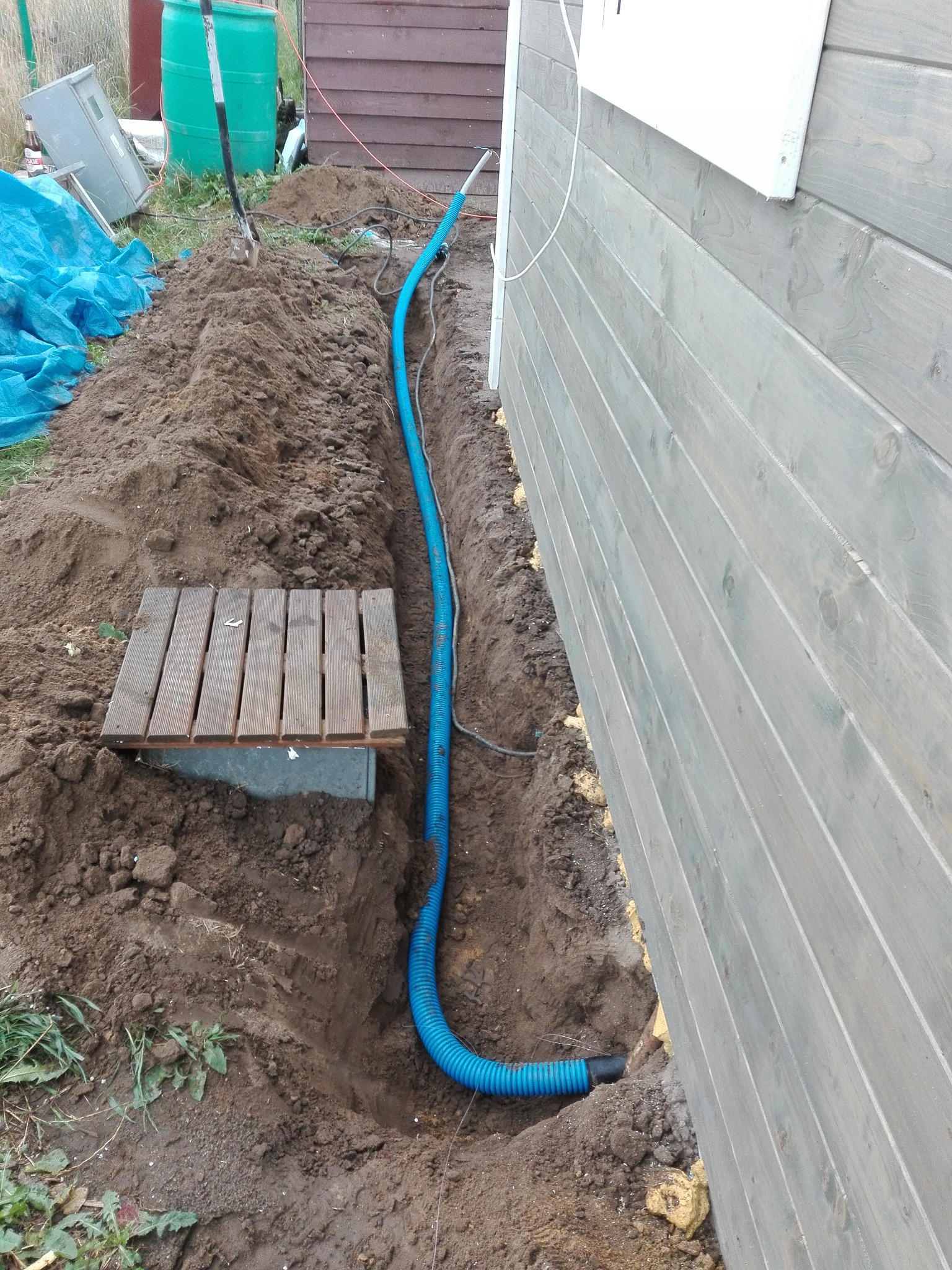 Wkopana niebieska rura karbowana biegnąca wzdłuż ściany domu z drewnianą elewacją, częściowo przykryta ziemią, obok wykopu widoczny drewniany podest.