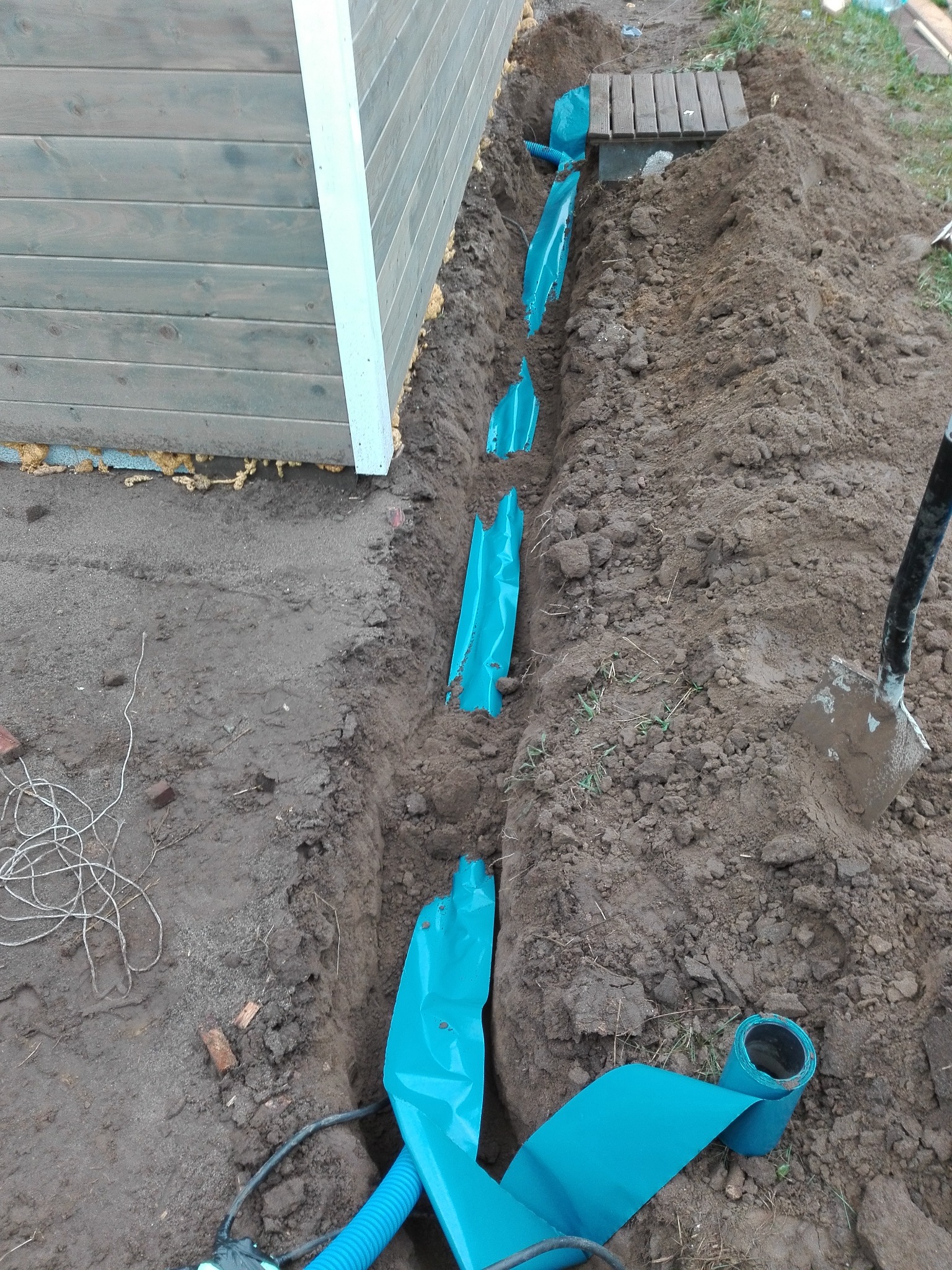 Wykop w ziemi z ułożoną niebieską folią ochronną, widoczna rura karbowana oraz szpadel wbity w ziemię obok