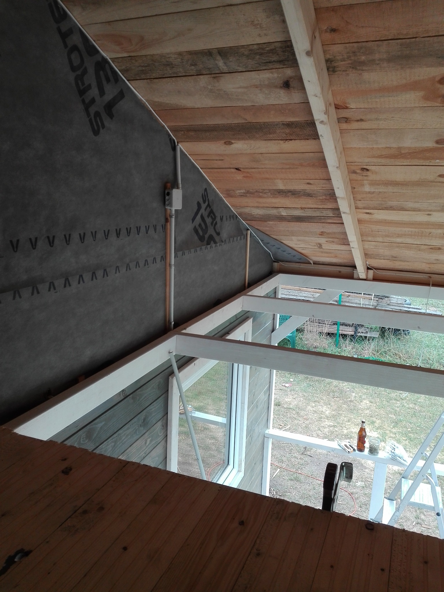 Wnętrze w trakcie budowy, widok na drewniany strop i ścianę z izolacją, widoczne okno z białymi ramami, na parapecie butelka, słoik i zszywacz tapicerski.