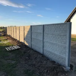 Długi, betonowy płot panelowy, wzniesiony wzdłuż granicy działki, z widoczną paletą i zarysem osoby w tle, na tle domu z białymi ścianami i błękitnego nieba.