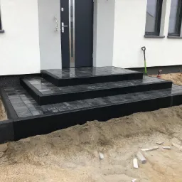 Nowoczesne, ciemnoszare schody wejściowe z kostki brukowej prowadzące do domu, widoczne narzędzia i niedokończone prace ziemne wokół, w tle biała elewacja budynku.