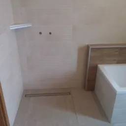 Wykończona łazienka z beżowymi płytkami, minimalistyczną półką i odpływem liniowym w podłodze, widoczna biała wanna z obudową imitującą drewno.