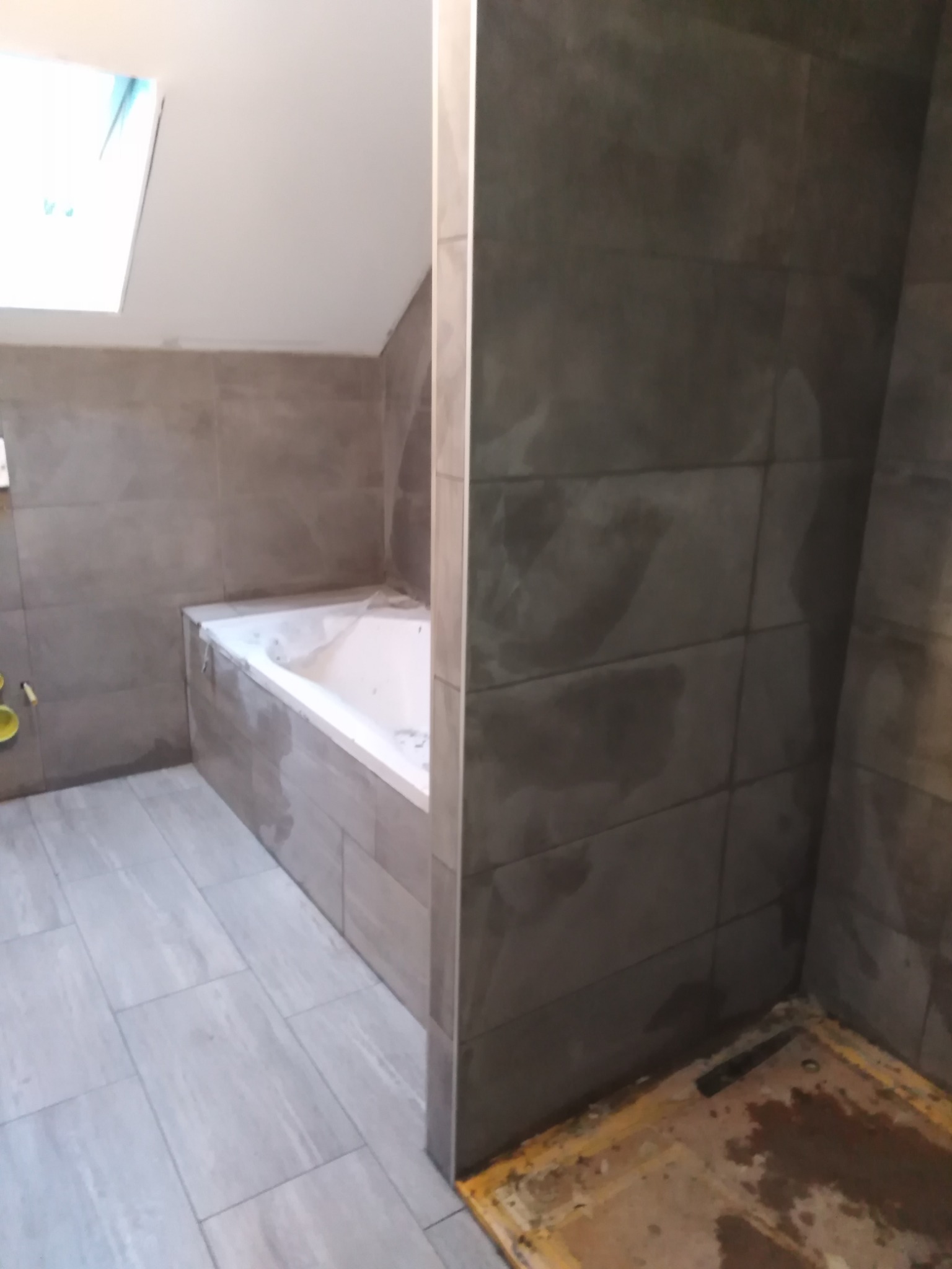 Częściowo wykończona łazienka z szarymi płytkami na ścianach i podłodze, widoczna wanna pod oknem dachowym i niezabezpieczony odpływ pod prysznicem.