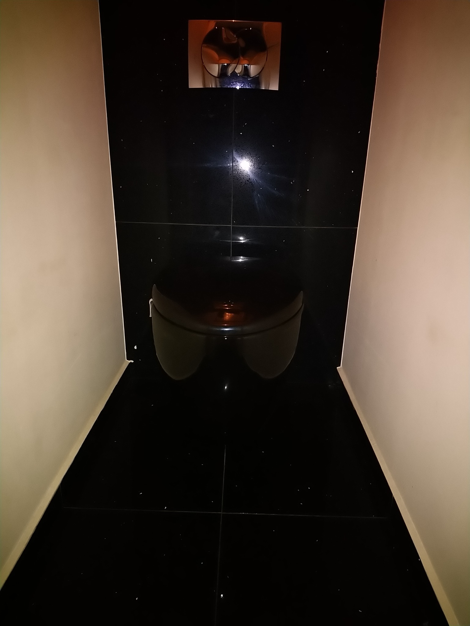 Czarna, błyszcząca toaleta podwieszana z chromowanym przyciskiem spłukiwania, otoczona kremowymi ścianami w małej wnęce wyłożonej czarnymi płytkami z delikatnym połyskiem.