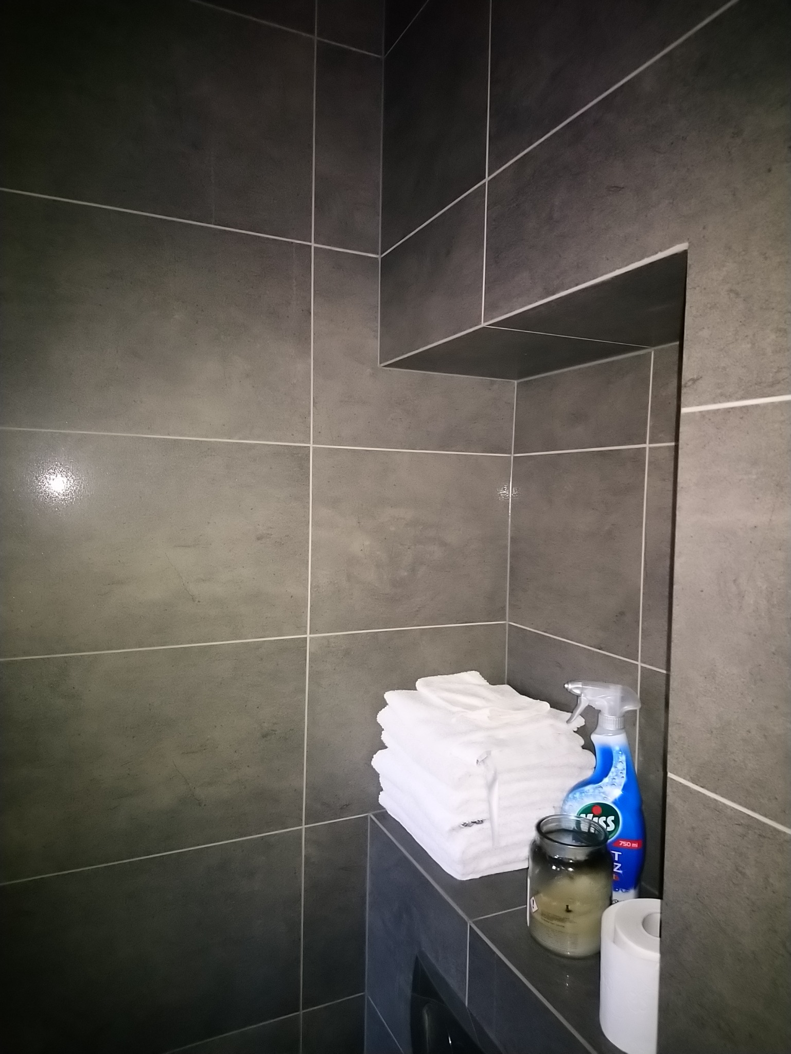 Szczegółowe ujęcie wykończonej łazienki w Szczecinie: ciemnoszare, matowe płytki wielkoformatowe z jasnymi fugami, stos białych ręczników, butelka płynu do czyszczenia i świeca w szklanym słoiku...