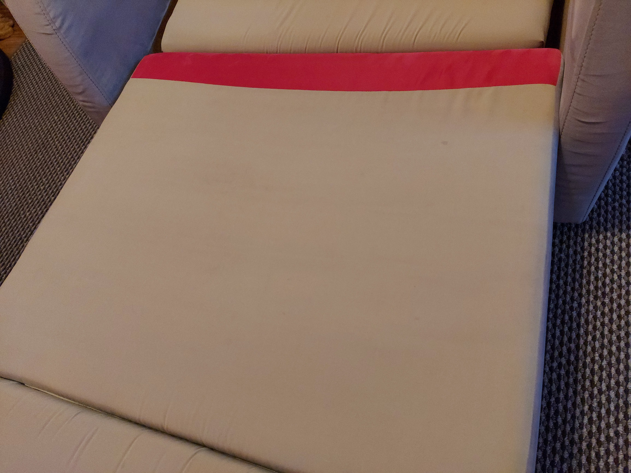 Rozłożona sofa z beżową tapicerką i czerwonym zagłówkiem, widoczna z góry na tle dywanu w geometryczny wzór.