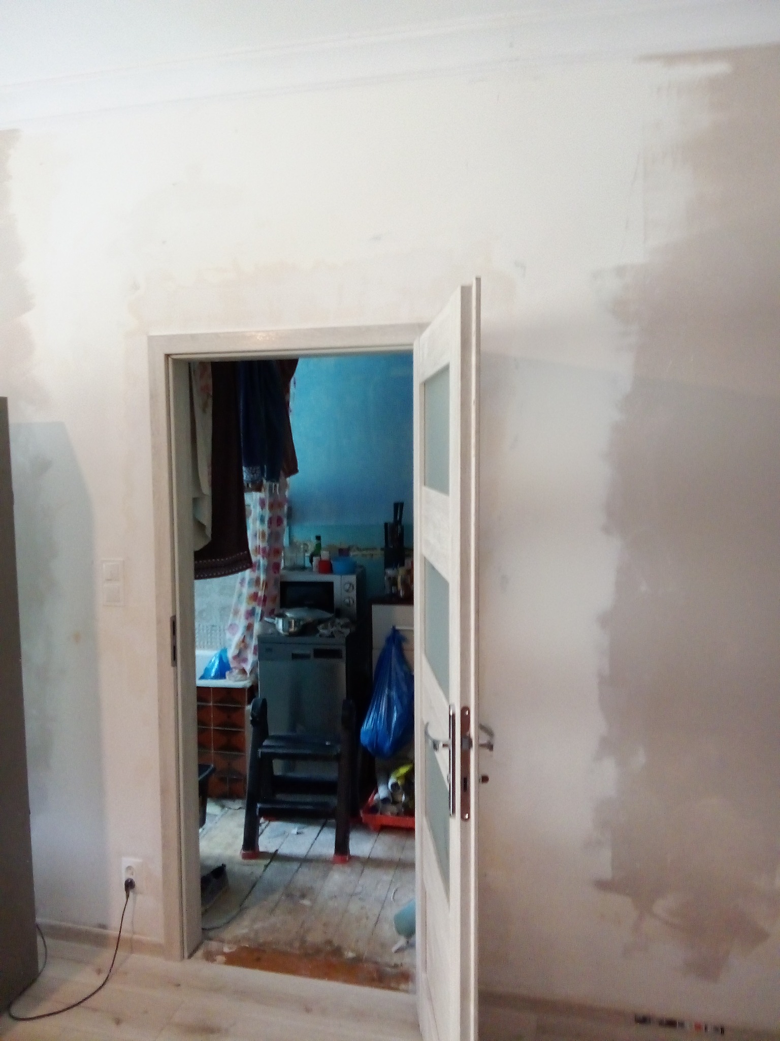 Ściana w trakcie prac wykończeniowych, widoczne ślady szpachlowania, otwarte drzwi do zagraconego pomieszczenia z widocznym wyposażeniem kuchni i łazienki.