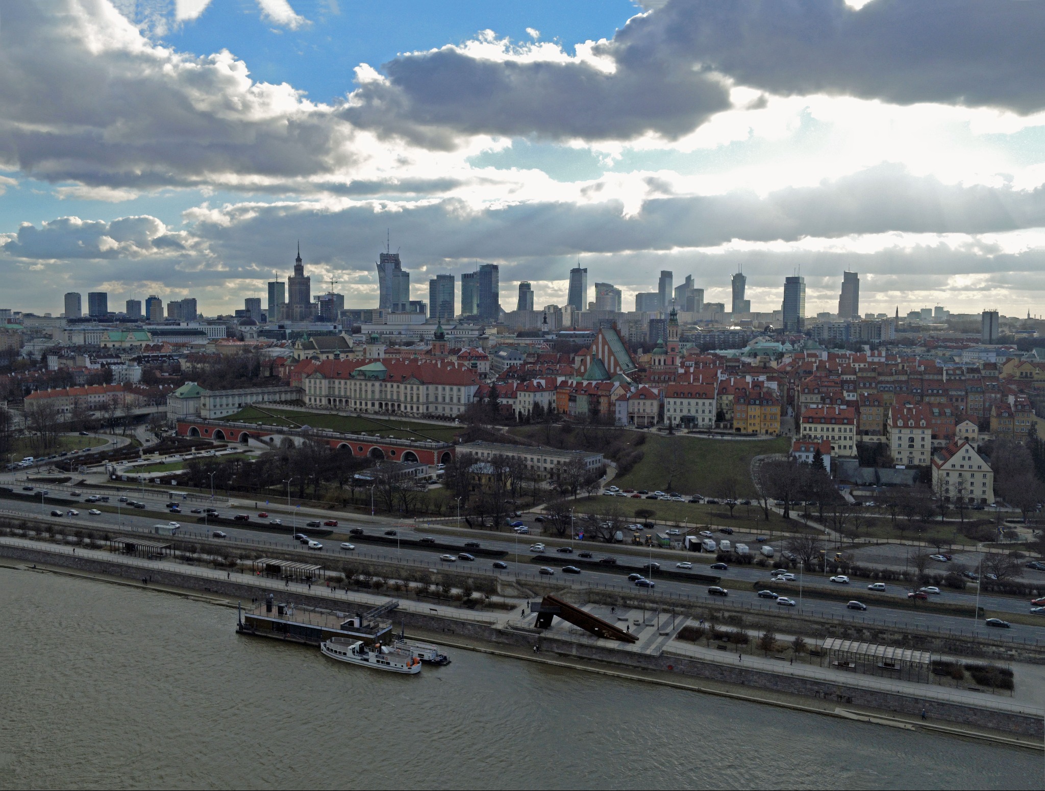 Panorama Warszawy z lotu ptaka, widok na Stare Miasto, Wisłę i nowoczesne wieżowce w tle, pod pochmurnym niebem z prześwitami słońca.