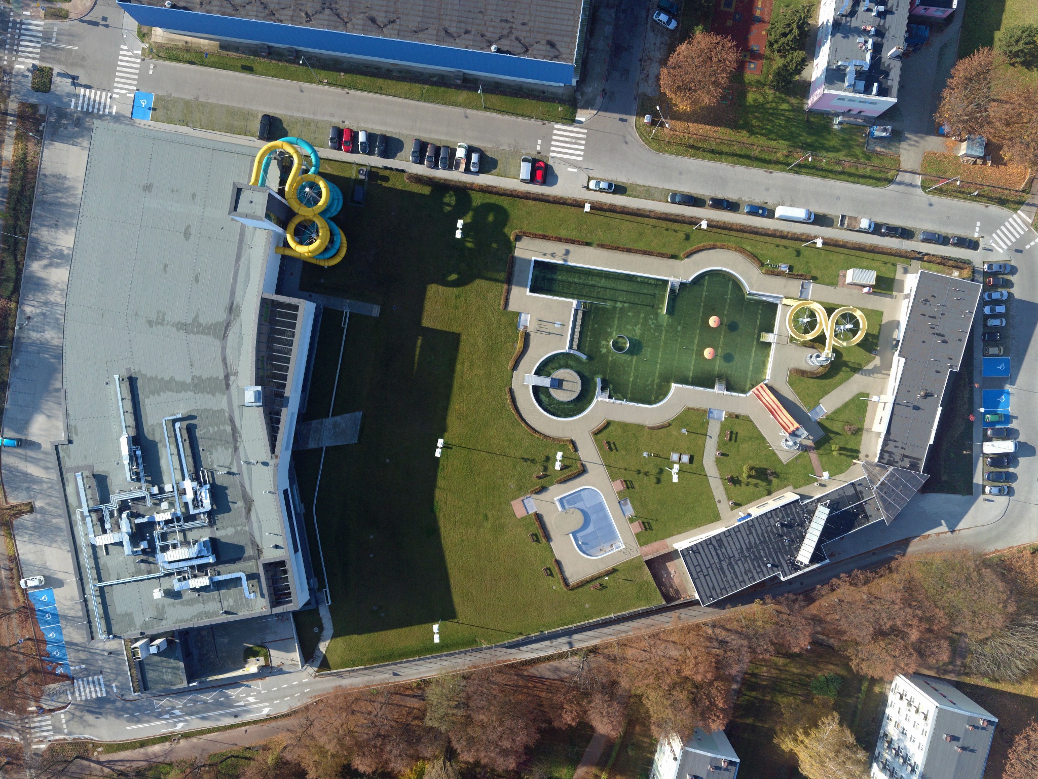 Widok z lotu ptaka na aquapark z kolorowymi zjeżdżalniami, basenami i zielonym terenem rekreacyjnym, otoczony parkingami i drzewami.