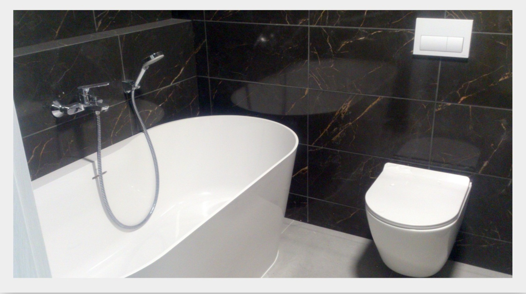 Nowoczesna łazienka z wolnostojącą białą wanną, chromowaną armaturą, podwieszaną toaletą i ciemnymi płytkami imitującymi marmur ze złotymi żyłami.