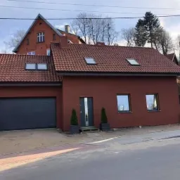 Ciemnoczerwony dom jednorodzinny z garażem i czerwonym dachem, widok z ulicy w słoneczny dzień.