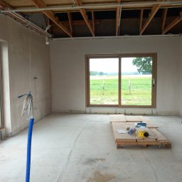 Rafal Kuriata - Wnętrze pomieszczenia w trakcie wykańczania z gładkimi, jasnymi ścianami, widocznym drewnianym stropem i instalacjami. Duże okno z drewnianą ramą otwiera widok na zielone pola. Na betonowej...