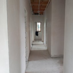 Rafal Kuriata - Wąski korytarz w trakcie prac wykończeniowych, z gładkimi, świeżo otynkowanymi ścianami i widocznymi rurami instalacji elektrycznej na drewnianym stropie. Na końcu korytarza okno i drabina.