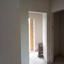 Rafal Kuriata - Wnętrze w trakcie budowy z gładkimi, jasnymi ścianami po tynkowaniu. Widoczne dwa otwory drzwiowe, surowa betonowa podłoga, zwisające okablowanie i paleta przy otworze zabezpieczonym płytą OSB.