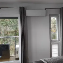 Biały klimatyzator ścienny zamontowany nad oknem z widokiem na zalesiony balkon, szare zasłony, nowoczesna lampa sufitowa.