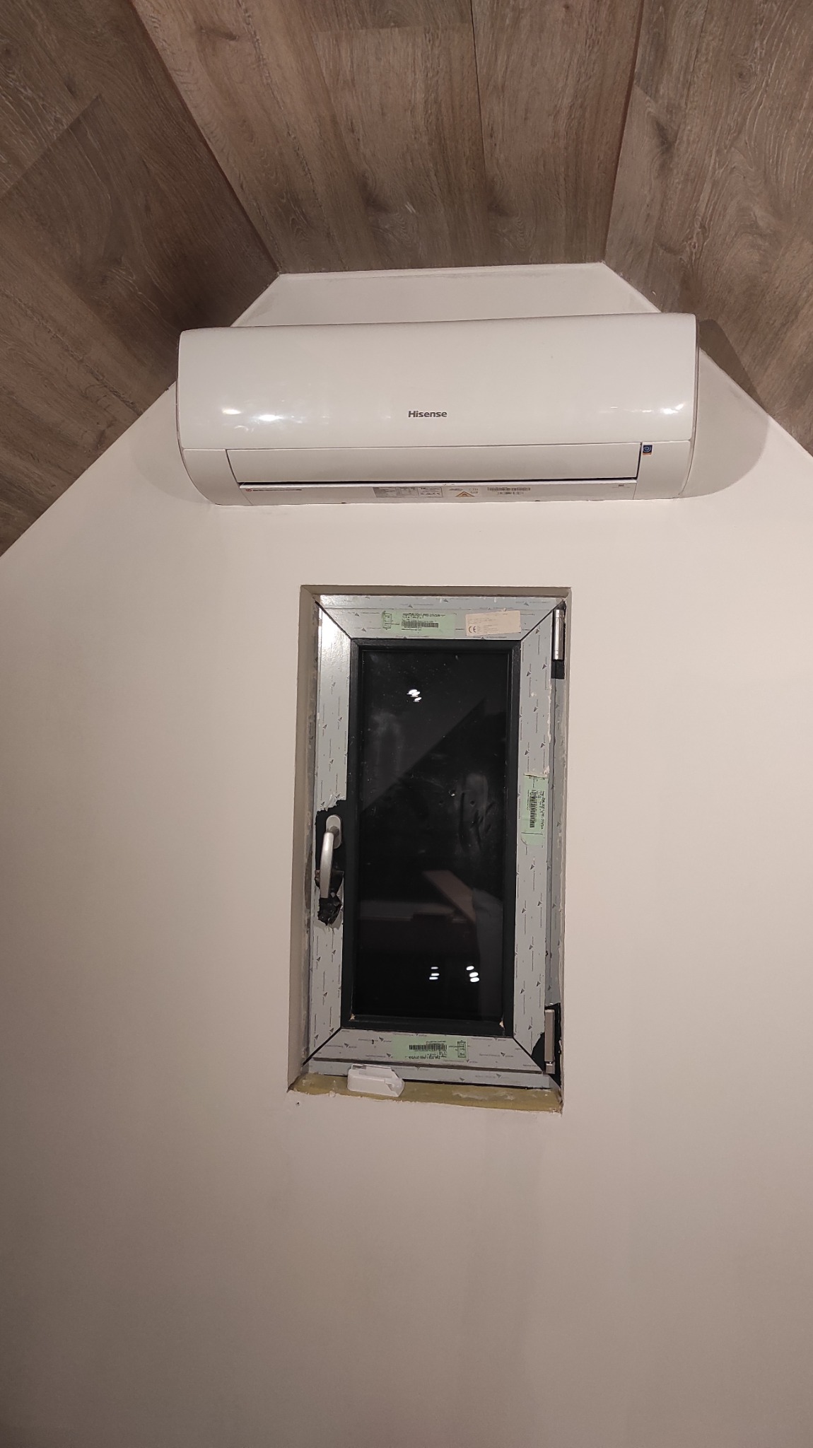 Biały klimatyzator marki Hisense zamontowany nad oknem z szarą ramą, widoczne naklejki na ramie okiennej, ściana pomalowana na biało, podłoga wykończona panelami imitującymi drewno.