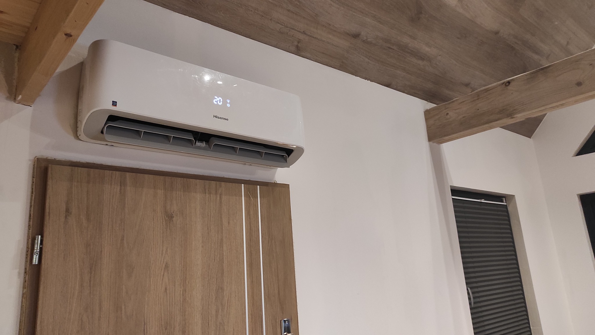 Biały klimatyzator Hisense zamontowany nad drewnianymi drzwiami w jasnym pomieszczeniu z drewnianymi belkami stropowymi.