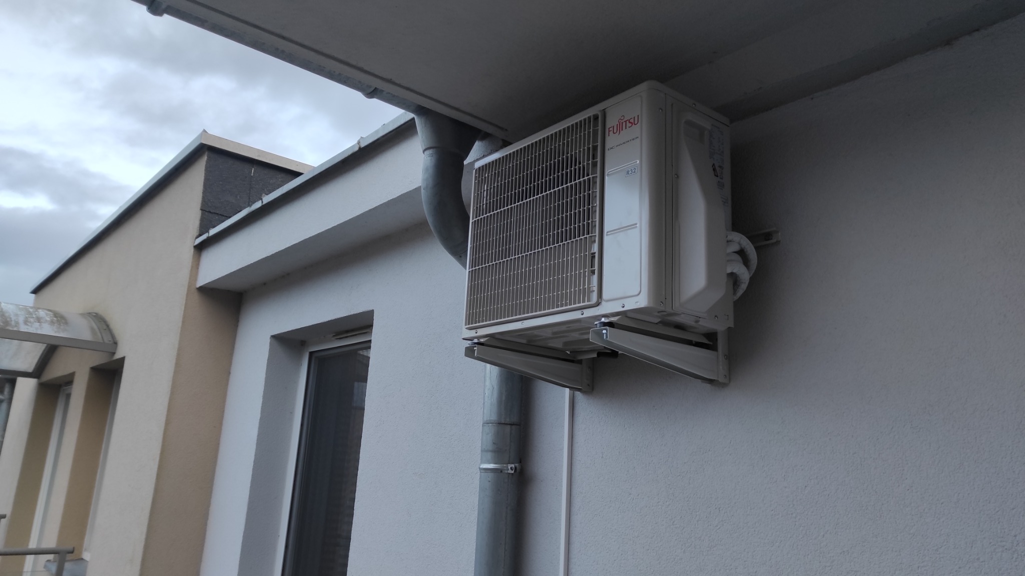 Zewnętrzna jednostka klimatyzatora Fujitsu zamontowana na ścianie budynku, widoczny przewód odprowadzający skropliny i rynna spustowa.