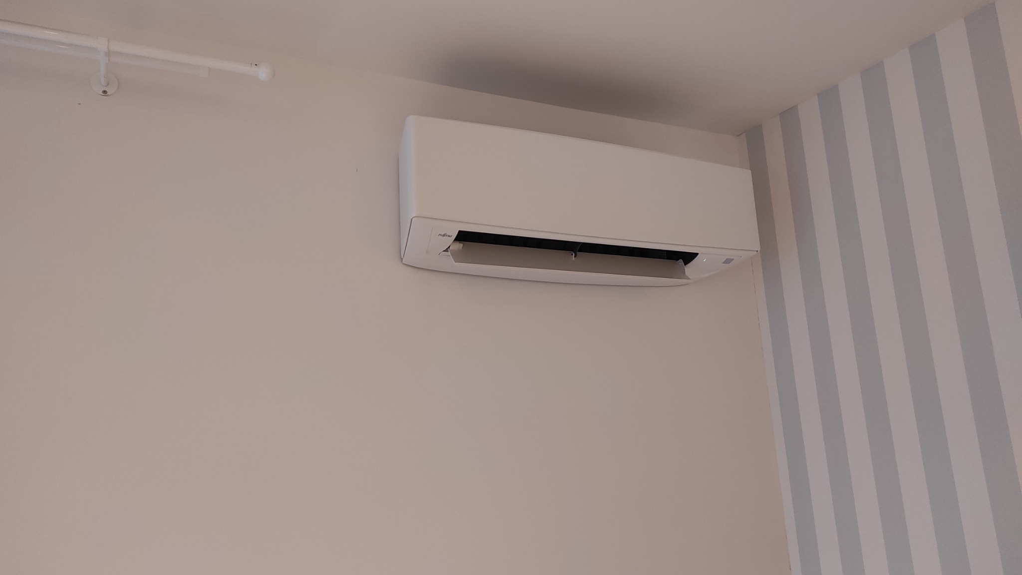 Biała jednostka wewnętrzna klimatyzacji Fujitsu zamontowana na ścianie w pokoju z tapetą w pionowe, szaro-białe pasy.