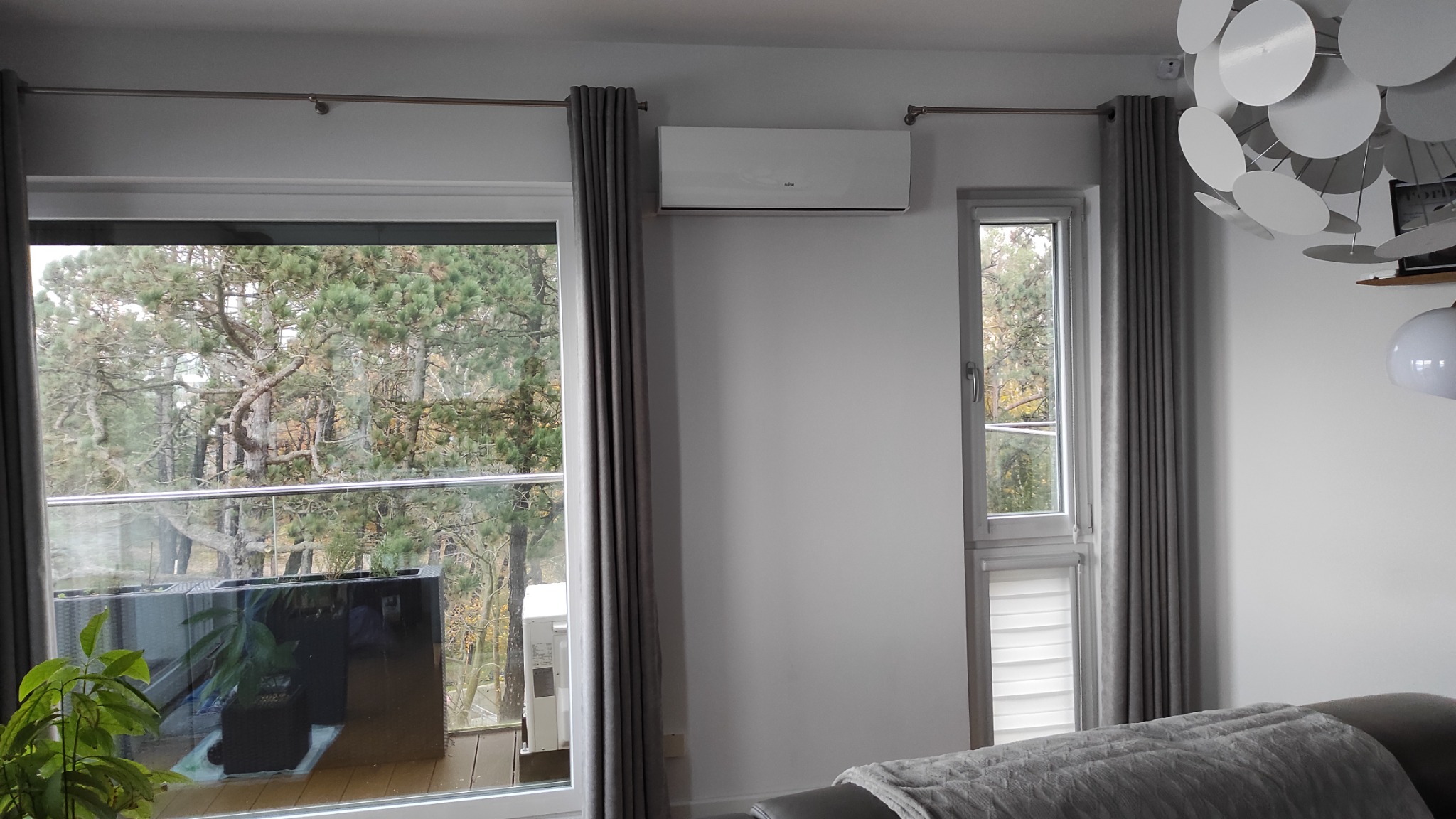 Biały klimatyzator ścienny zamontowany nad oknem z widokiem na zalesiony balkon, szare zasłony, nowoczesna lampa sufitowa.