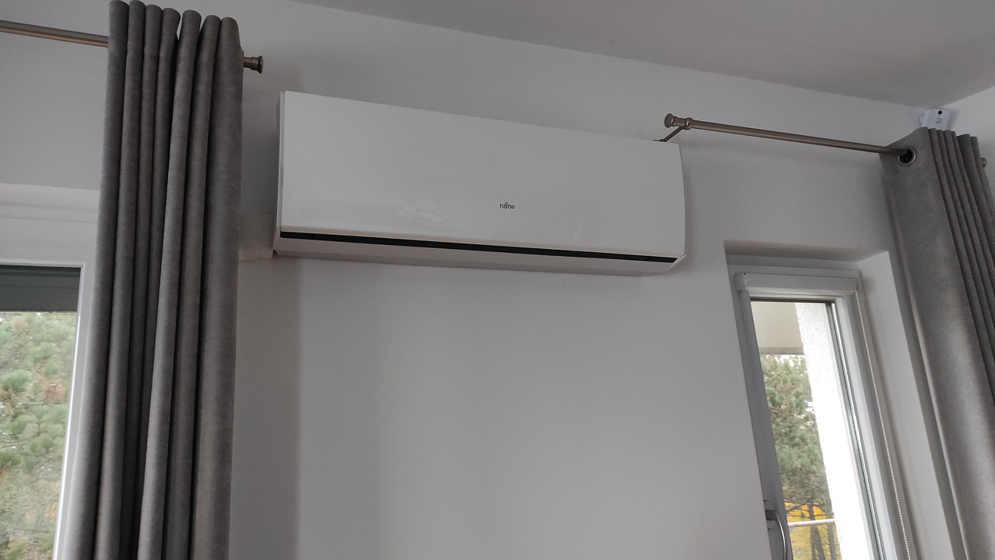 Biały klimatyzator Fujitsu zamontowany na ścianie w jasnym pokoju z szarymi zasłonami i widokiem na drzewa za oknem.