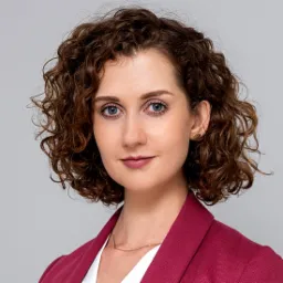 Portret kobiety z kręconymi włosami, błękitnymi oczami i w bordowej marynarce, uśmiechająca się delikatnie do kamery na szarym tle.