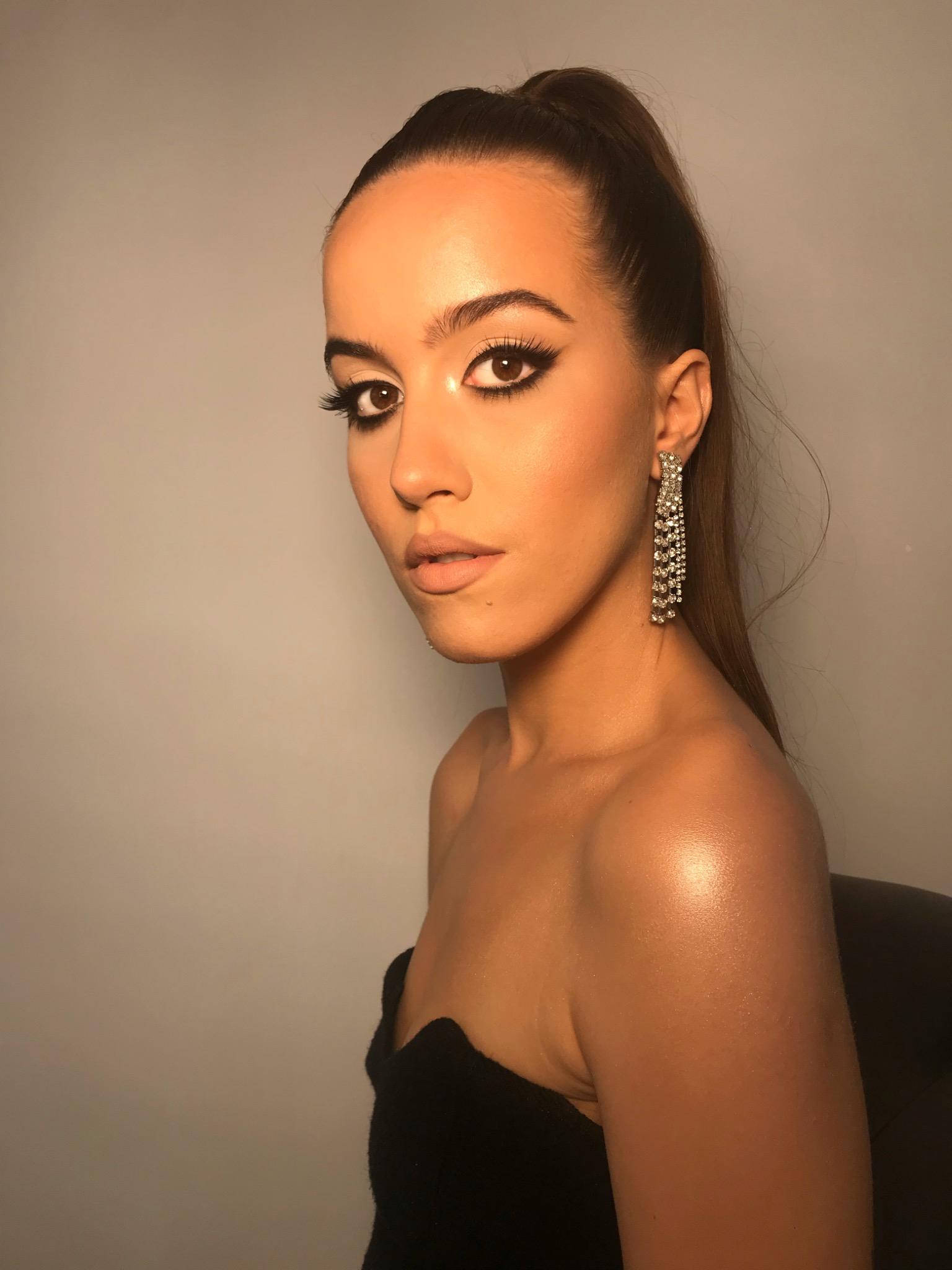 Portret kobiety z wyrazistym makijażem oczu w stylu cat-eye, błyszczącą skórą i długimi, wiszącymi kolczykami z kryształków, włosy upięte w wysoki kucyk.