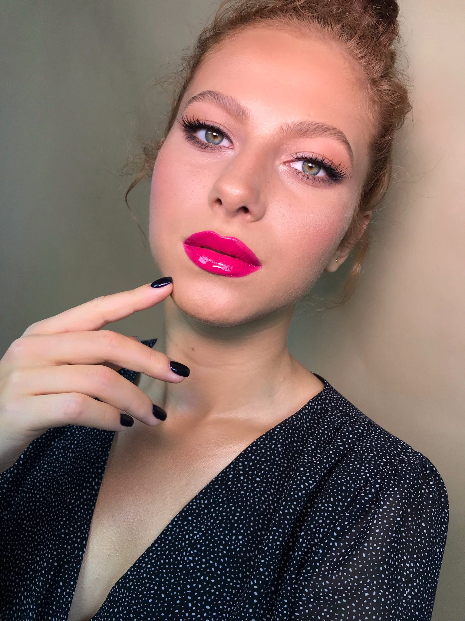 Portret kobiety z intensywnym różowym makijażem ust i ciemnym manicure, dłoń dotyka policzka, wzrok skierowany w górę, na beżowym tle.