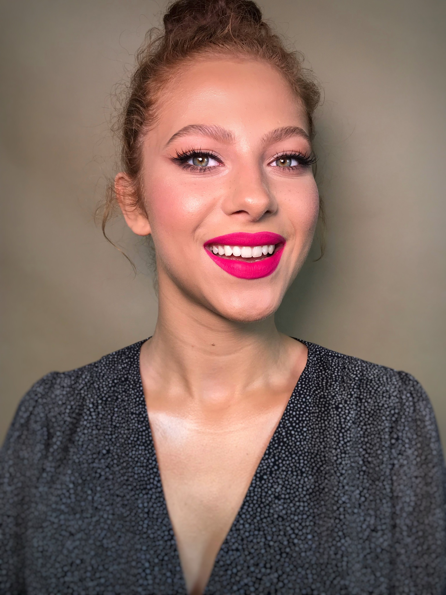 Uśmiechnięta kobieta z intensywnym różowym odcieniem szminki, podkreślonymi oczami z wydłużonymi rzęsami i delikatnym różem na policzkach, włosy upięte w kok, ubrana w bluzkę w drobne kropki.