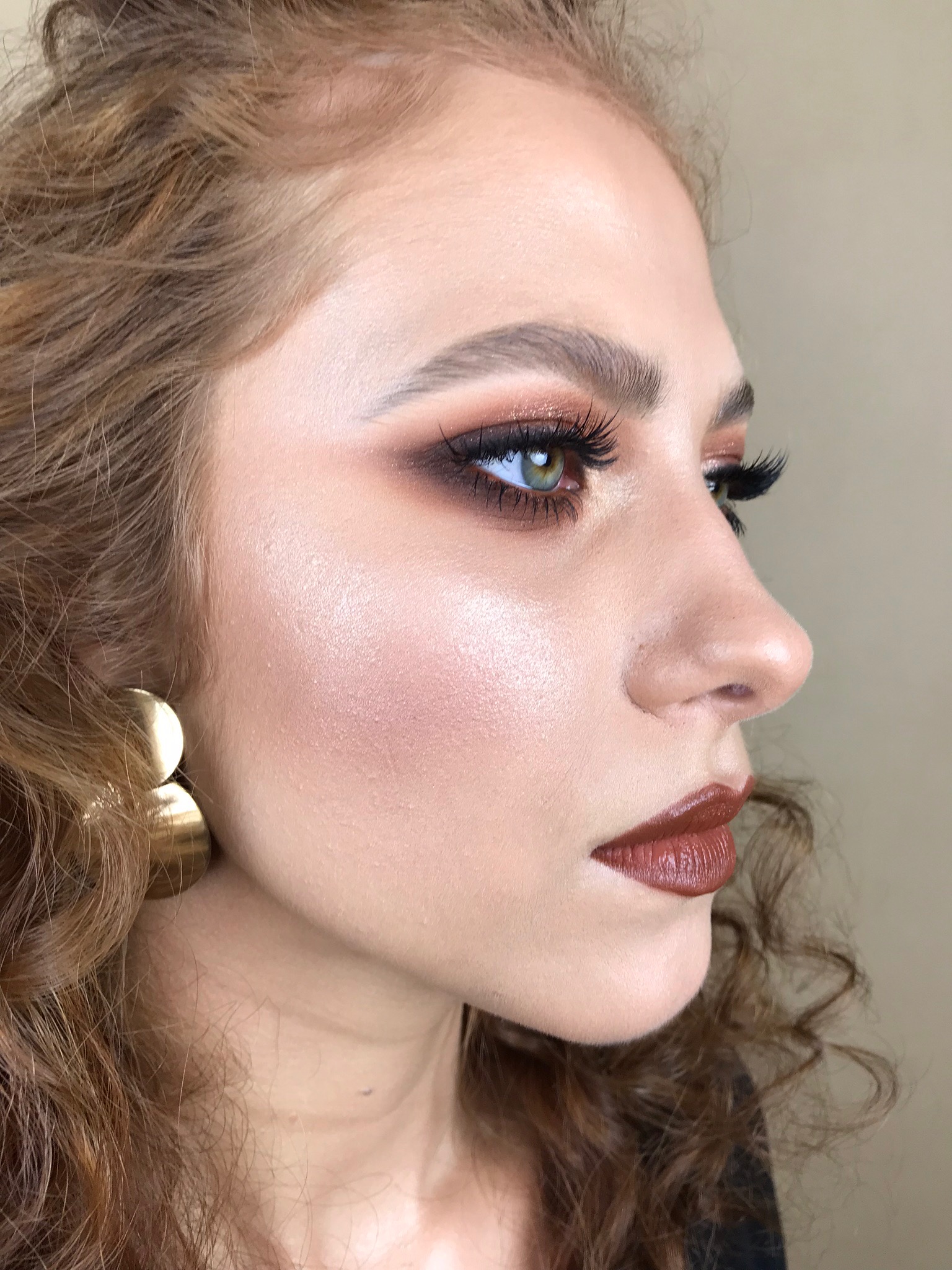 Portret z profilu kobiety z profesjonalnym makijażem oczu w odcieniach brązu i błyszczącym rozświetlaczem na policzkach, podkreślone usta w kolorze brązowym, duże złote kolczyki.
