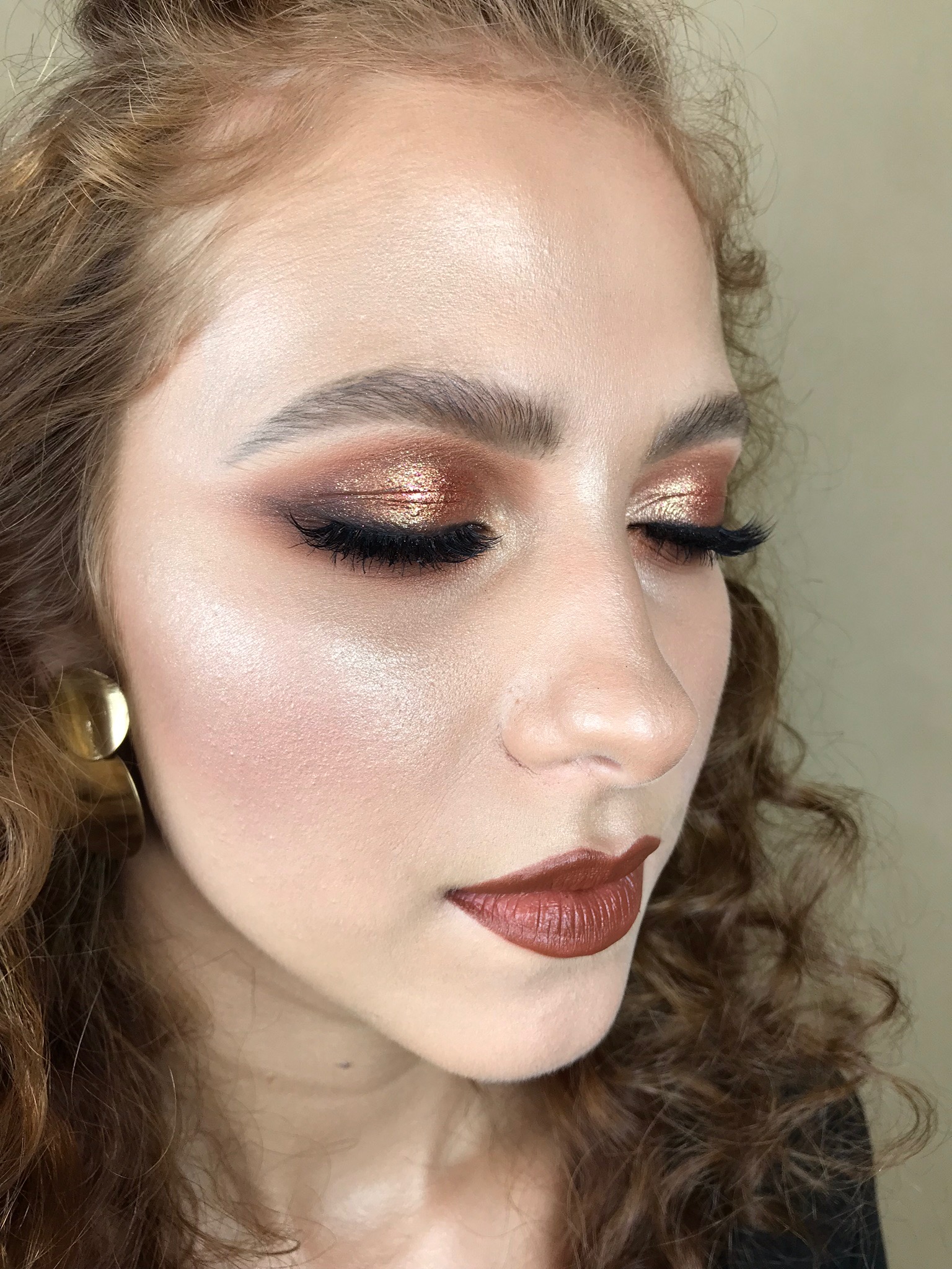 Portret kobiety z zamkniętymi oczami, prezentujący intensywny, złoto-brązowy makijaż oczu z wyraźną kreską i sztucznymi rzęsami, podkreślone kości policzkowe z delikatnym rozświetlaczem oraz usta...